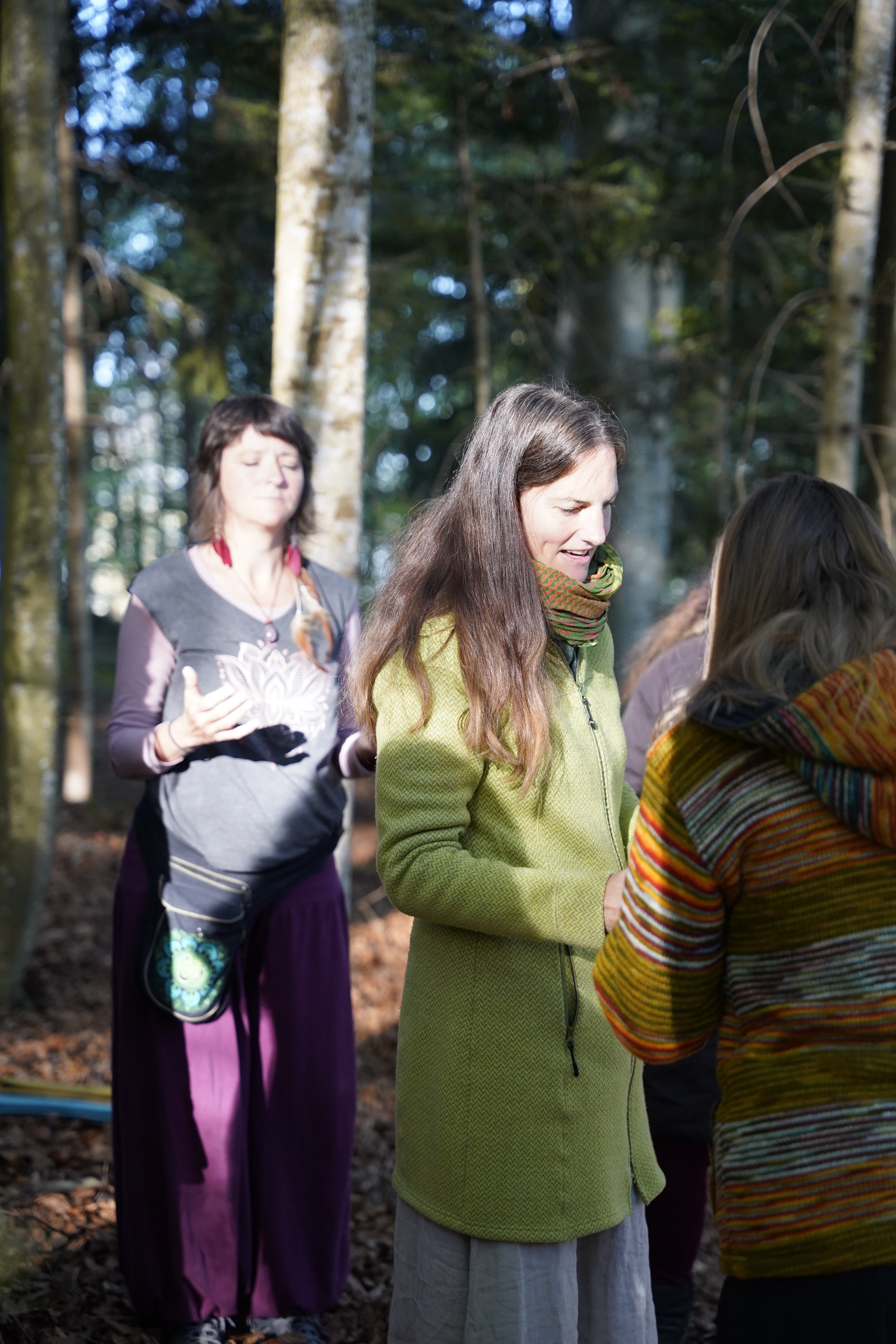 Drei Frauen in einer Waldumgebung, die miteinander sprechen, eine Person trägt eine grüne Jacke, eine andere trägt einen bunten Pullover, im Hintergrund steht eine weitere Frau mit Blick nach oben.