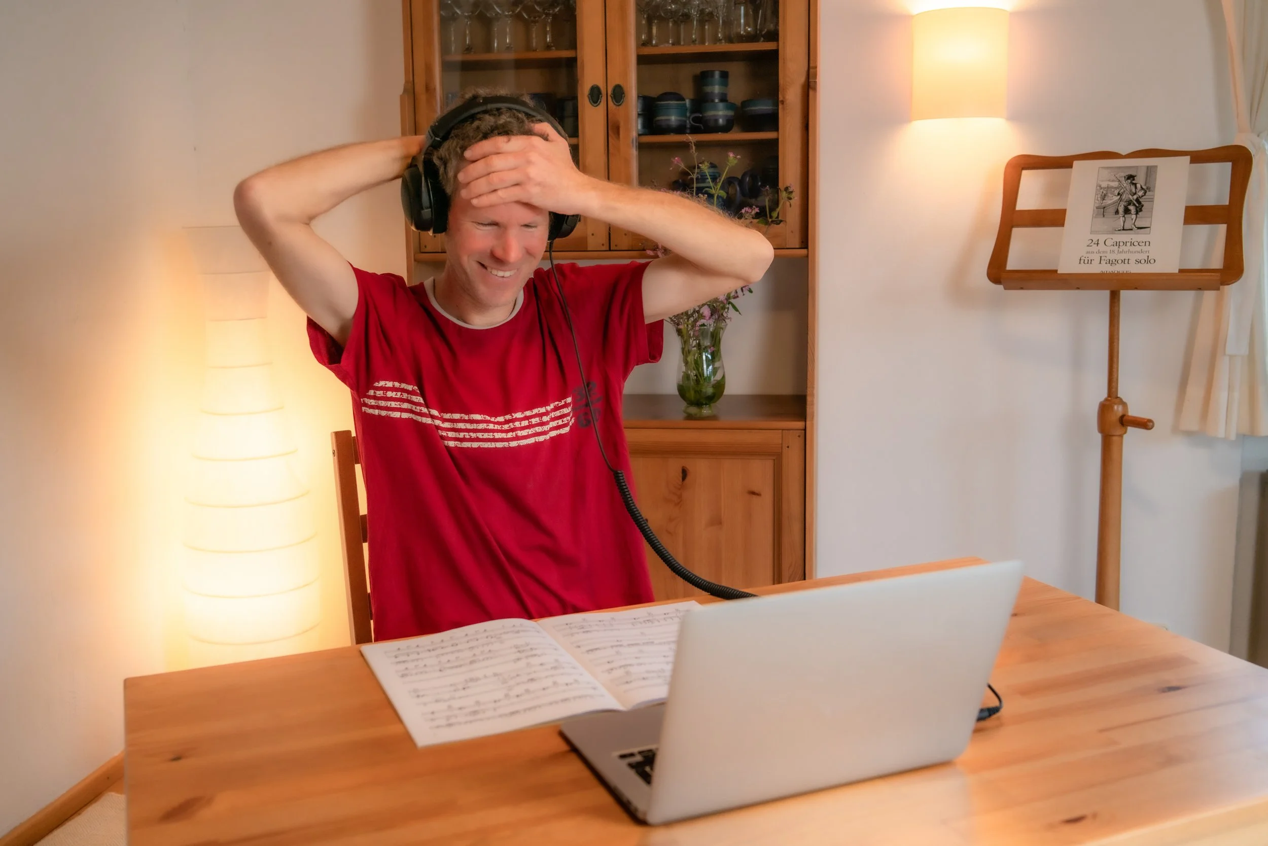 Junge Person sitzt an einem Holztisch, trägt Kopfhörer, lächelt und hat die Hände am Kopf. Im Vordergrund eine offene Noten- oder Notizbuch, im Hintergrund ein Schrank mit Gläsern und Geschirr, eine Vase mit Blumen und eine Stehlampe. Auf einem Holzständer steht eine gedruckte Note mit einem Musikerbild und Text.