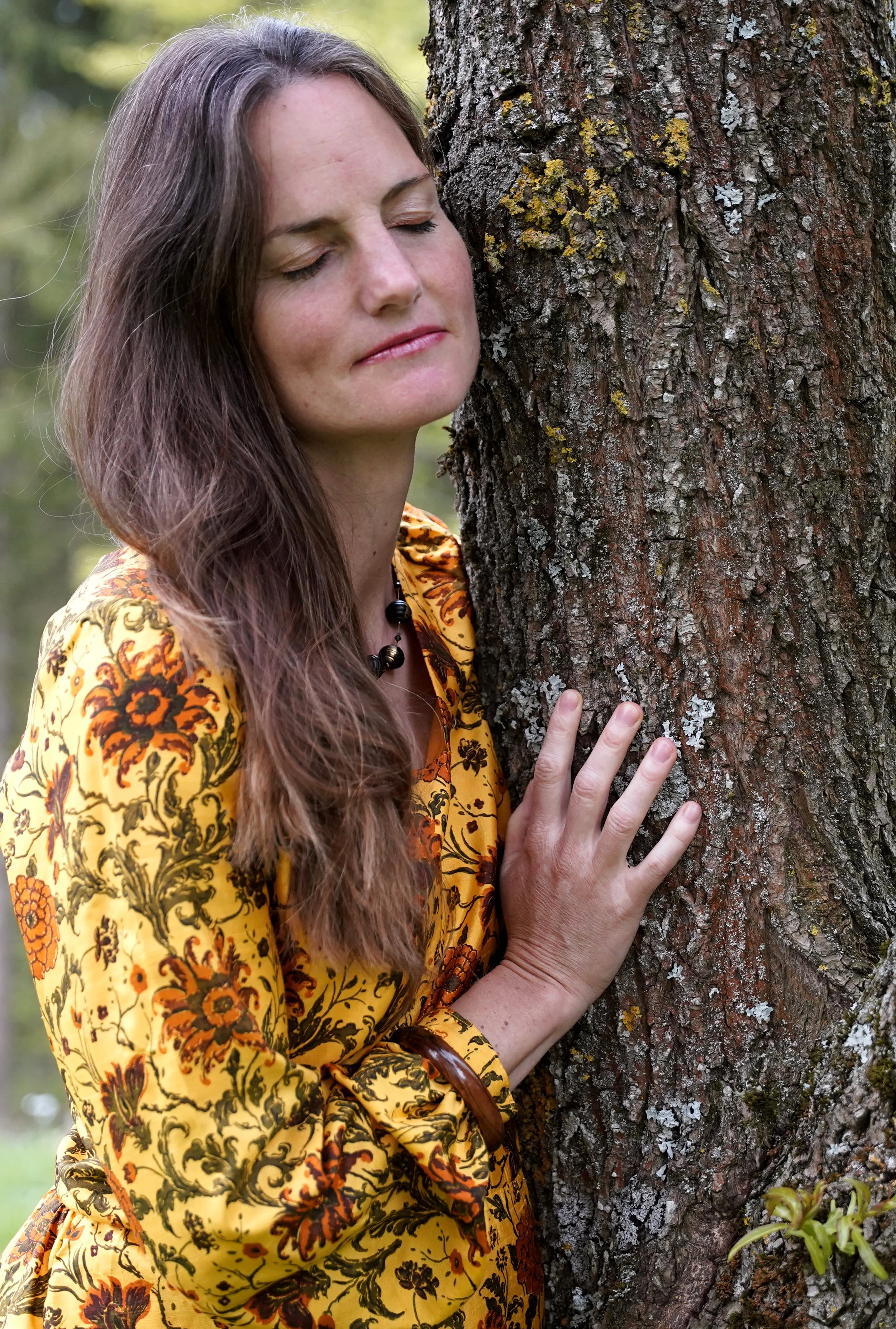 Frau mit langen braunen Haaren umarmt einen Baum mit geschlossener Augen.