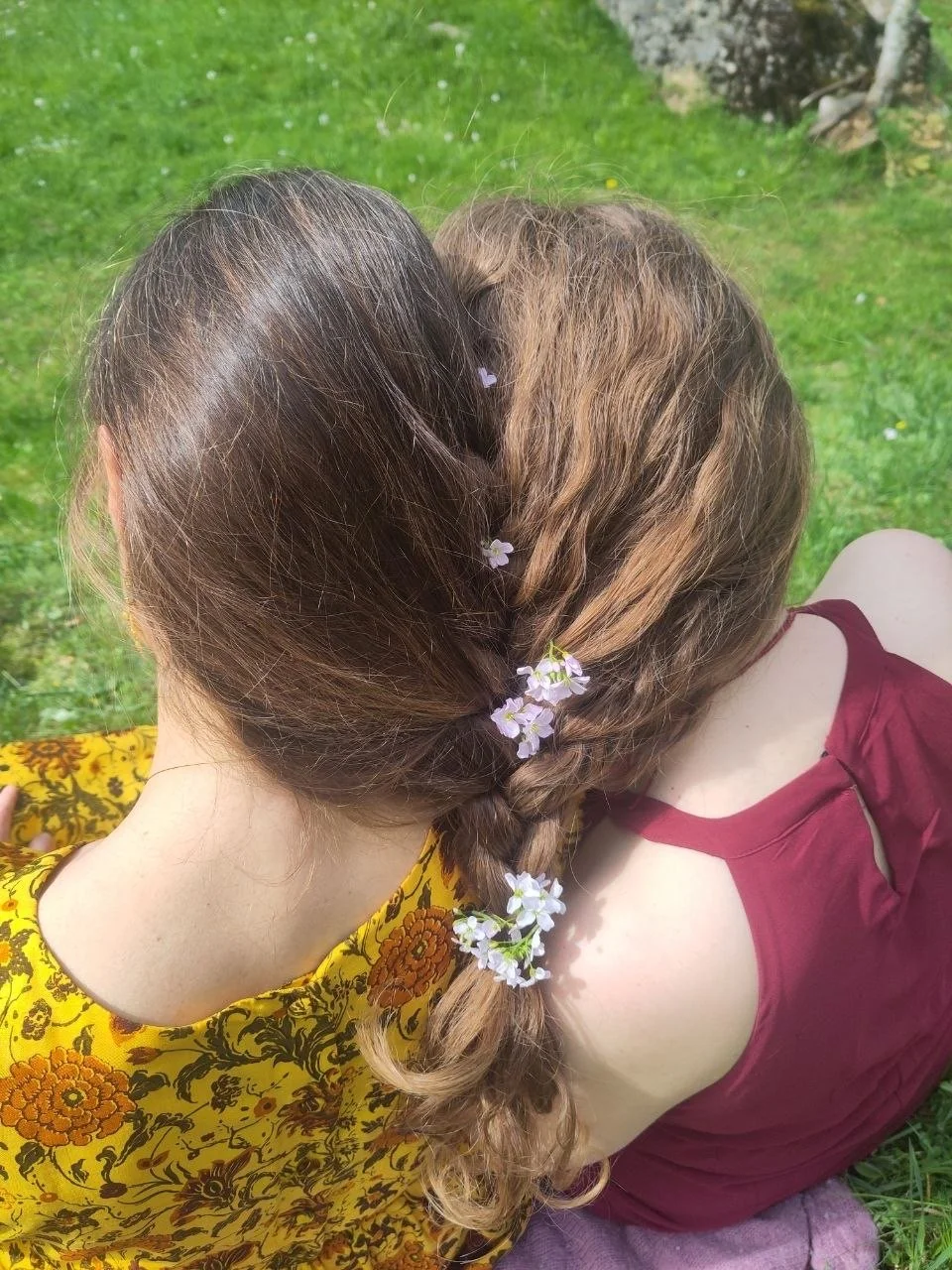 Zwei Frauen mit braunen, gewellten Haaren sitzen im Gras. Eine trägt eine gelbe Bluse mit orangfarbenen Blumen, die andere ein burgunderrotes Top. Die eine hat kleine weiße Blumen im Haar.