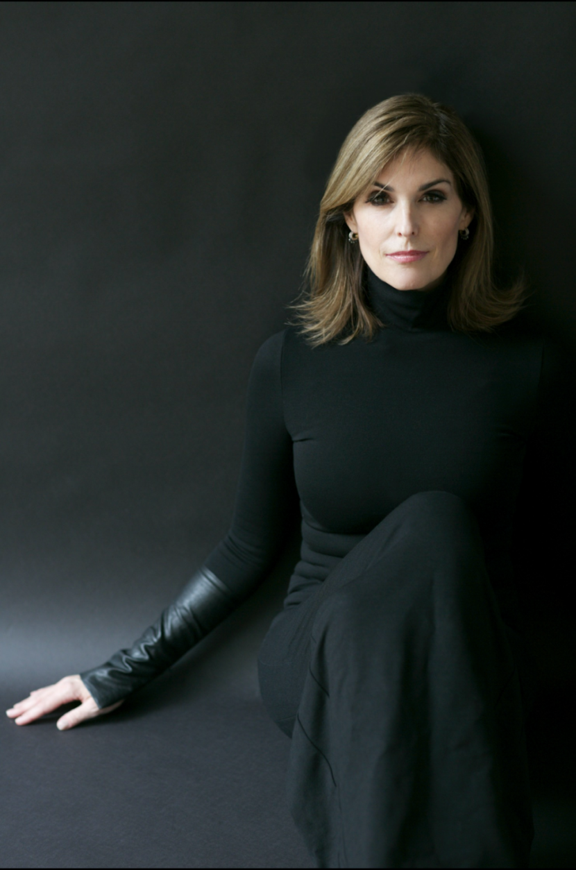 Lisa Pierpont with shoulder-length blonde hair, wearing a black turtleneck and long black gloves, sitting against a dark background.