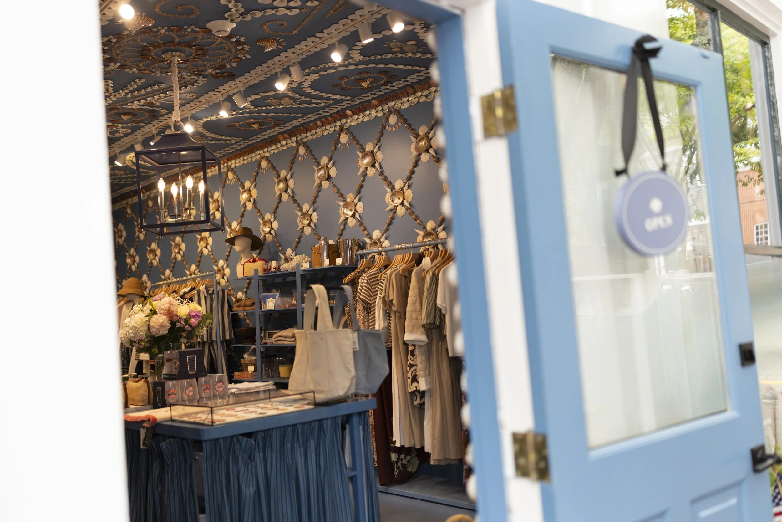 View of a clothing store interior seen through a partially open blue door with an 'Open' sign hanging. Inside, clothes are displayed on racks, and there is decorative wall art made of shells and a bouquet of flowers on a table.