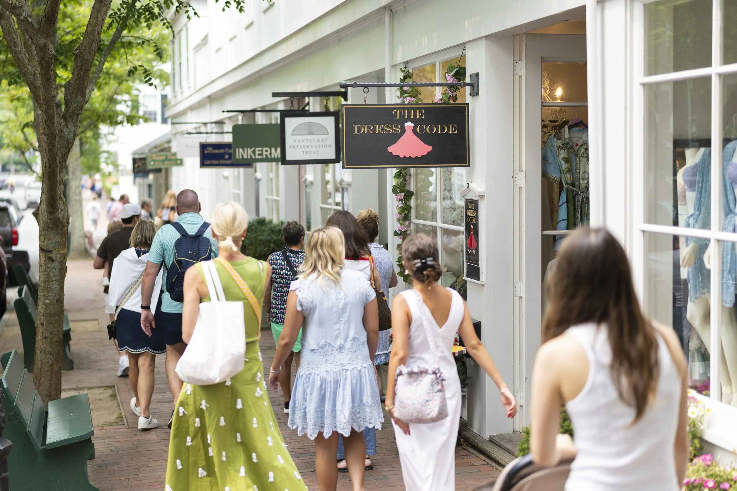 People walking along a sidewalk lined with shops, including one with a sign for 'The Dress Code' featuring a pink dress icon. Trees and greenery are visible, and some shop windows display clothing items.