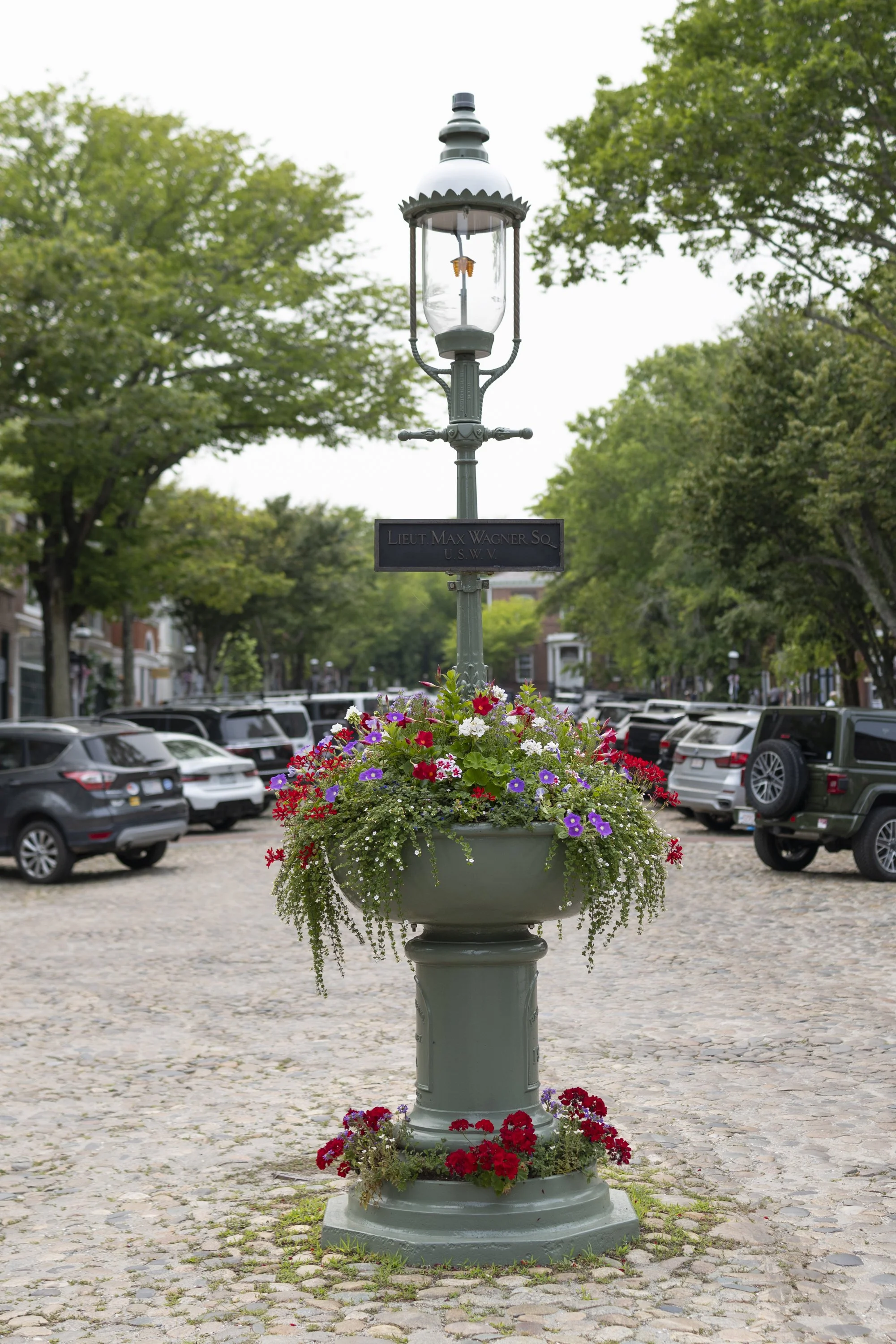 A decorative streetlamp and historic non-use fountain with a flower planter at its base, filled with colorful flowers, situated in a cobblestone plaza on Main Street with trees and parked cars in the background.