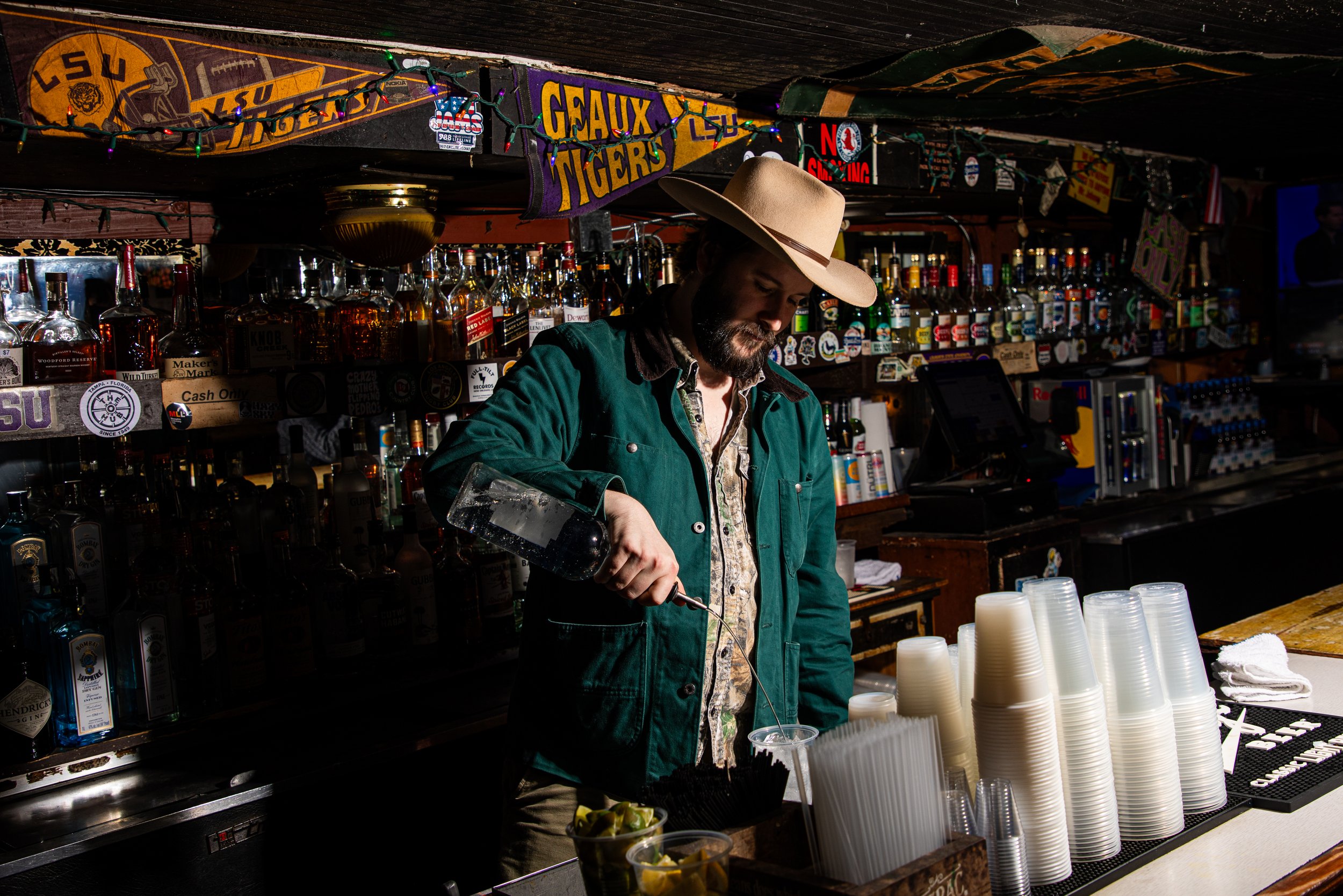 A man in a cowboy hat and green jacket standing behind a bar, pouring a drink into a plastic cup. The bar is stocked with various liquor bottles, and there are disposable cups stacked on the counter. The background has LSU Tigers and Geaux Tigers banners, along with string lights.