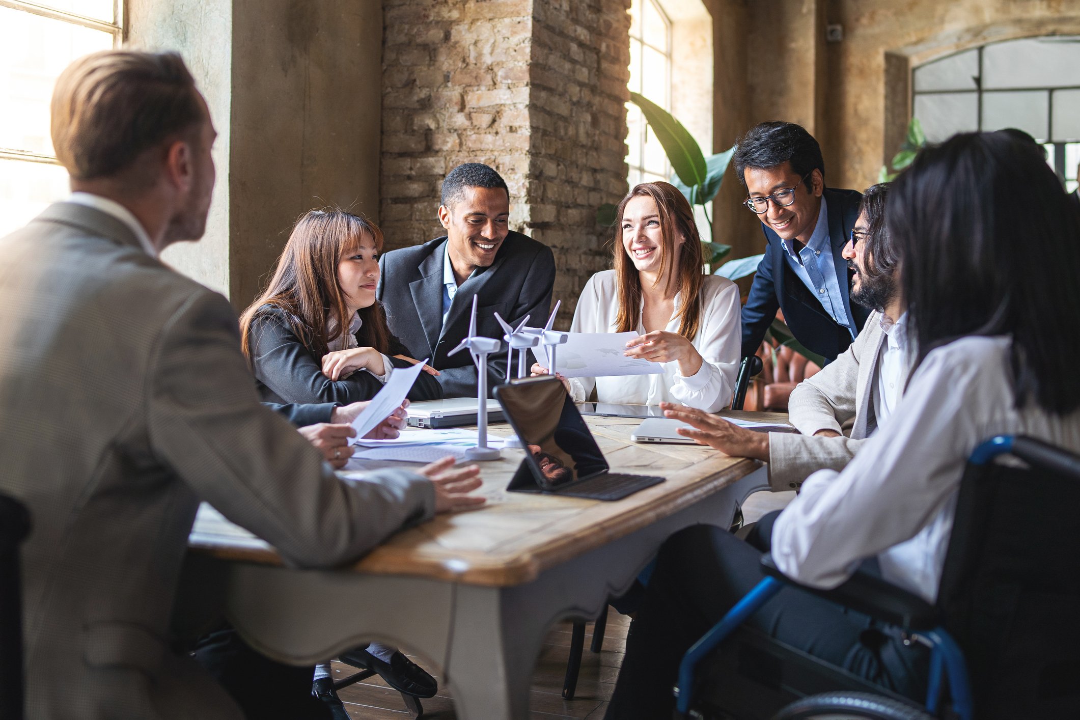 A diverse group of people in a business meeting, sitting around a wooden table in a brick-walled room with large windows, discussing and smiling. There are wind turbine models on the table.