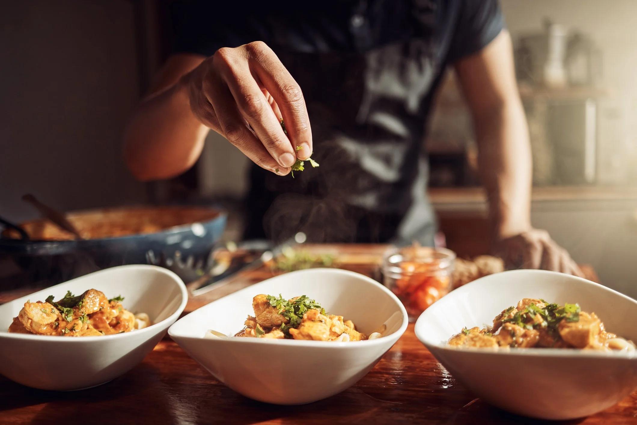 A person garnishes bowls of food with fresh herbs in a dimly lit kitchen.
