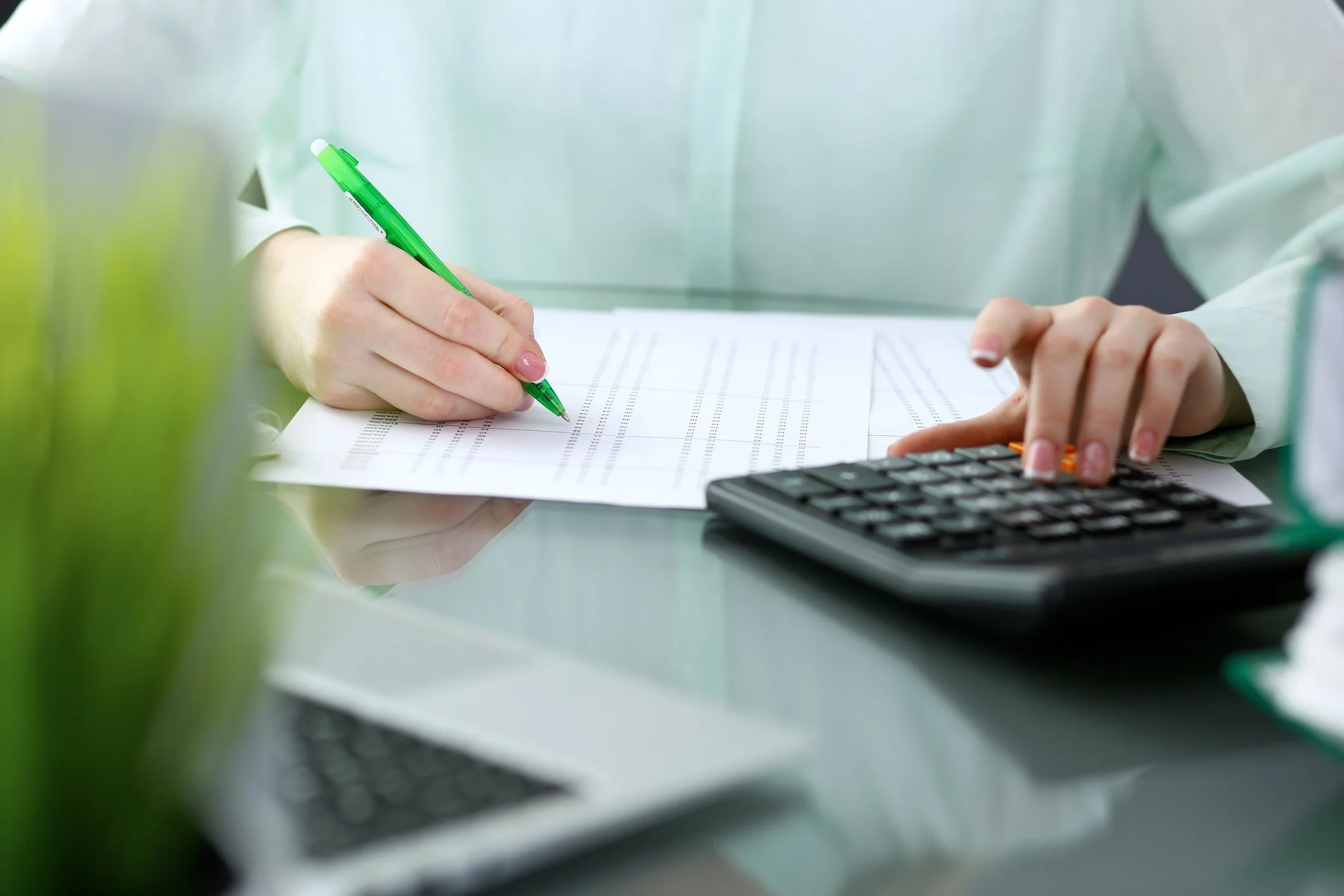 Close-up of a person's hands working with a calculator, a green pen, and document summaries on a desk, with blurred office background.