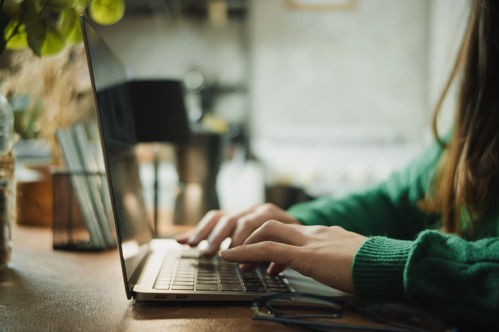 Person wearing a green sweater typing on a laptop keyboard with a blurred indoor background.