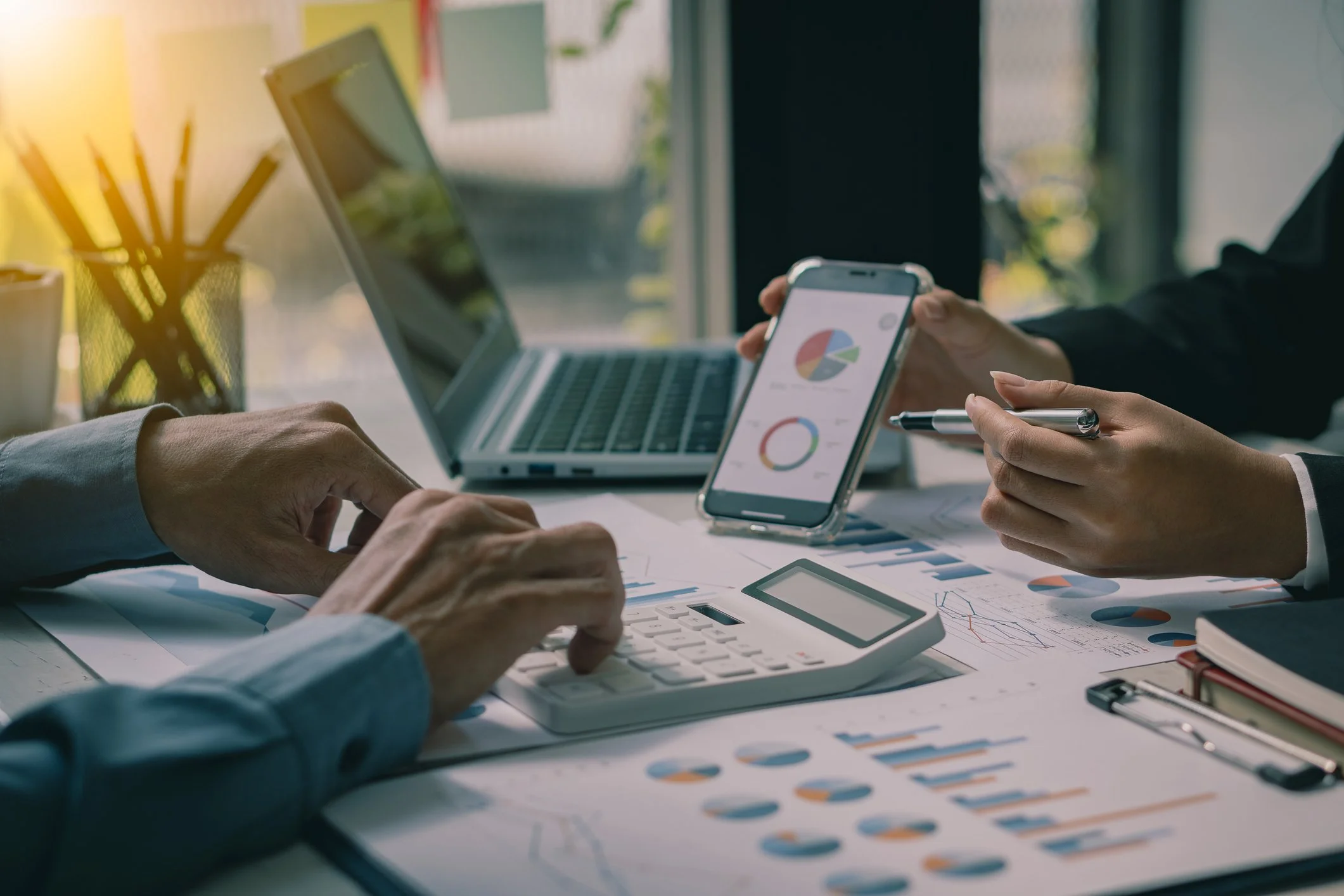 People working on financial charts and graphs with a laptop and smartphone on a desk in a well-lit office.