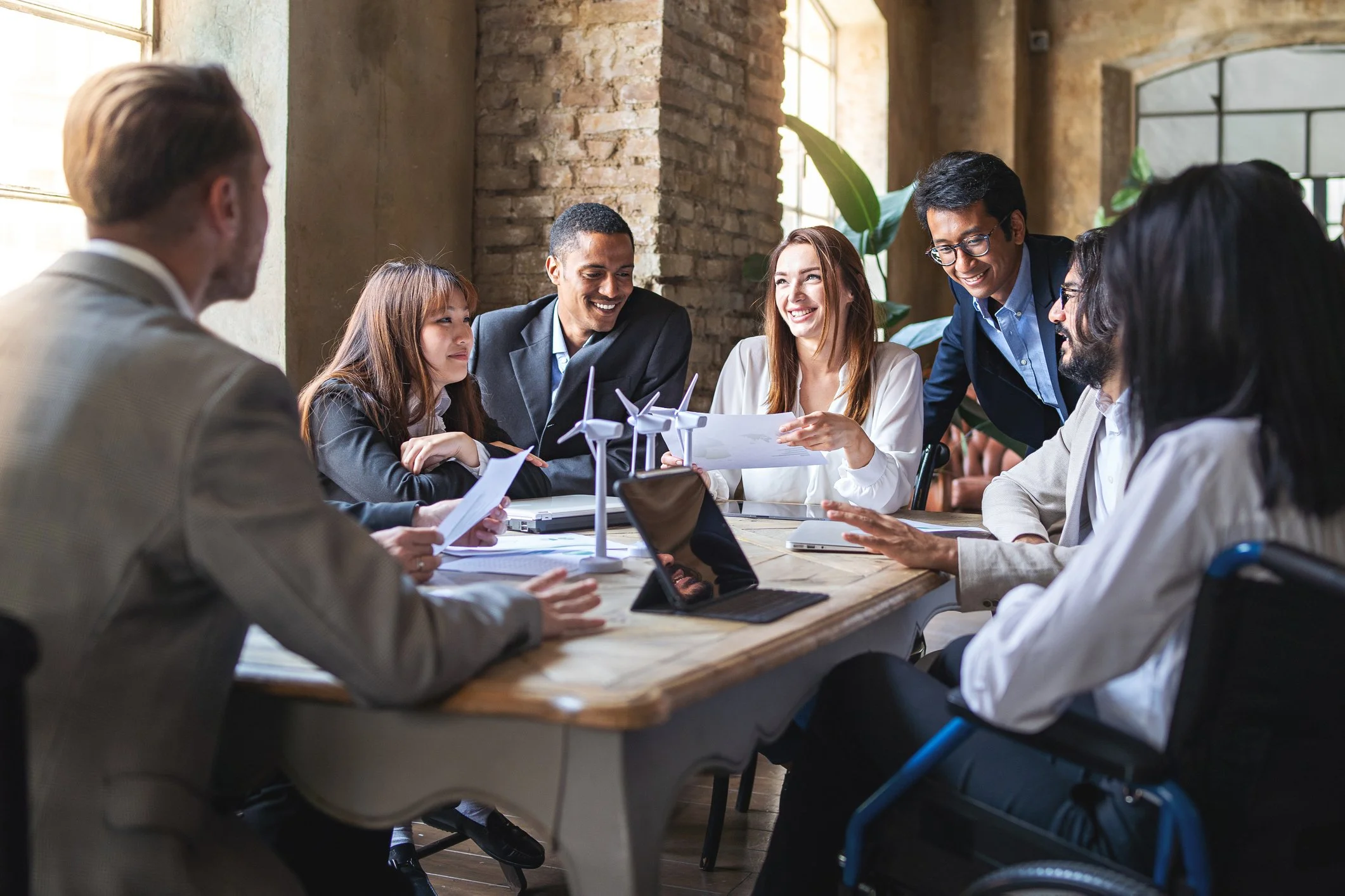 A diverse group of professionals gathered around a wooden table in a modern, industrial-style office with brick walls and large windows, discussing and reviewing documents and models, including miniature wind turbines.
