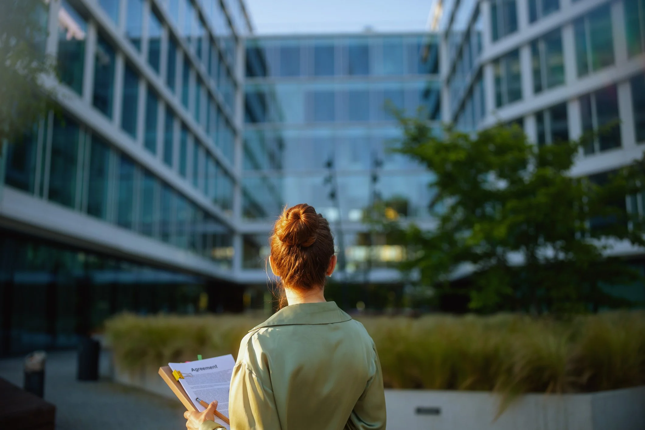 A person with red hair tied in a bun, holding a clipboard with a document titled 'Agreement,' standing outdoors in front of a modern glass building with trees.