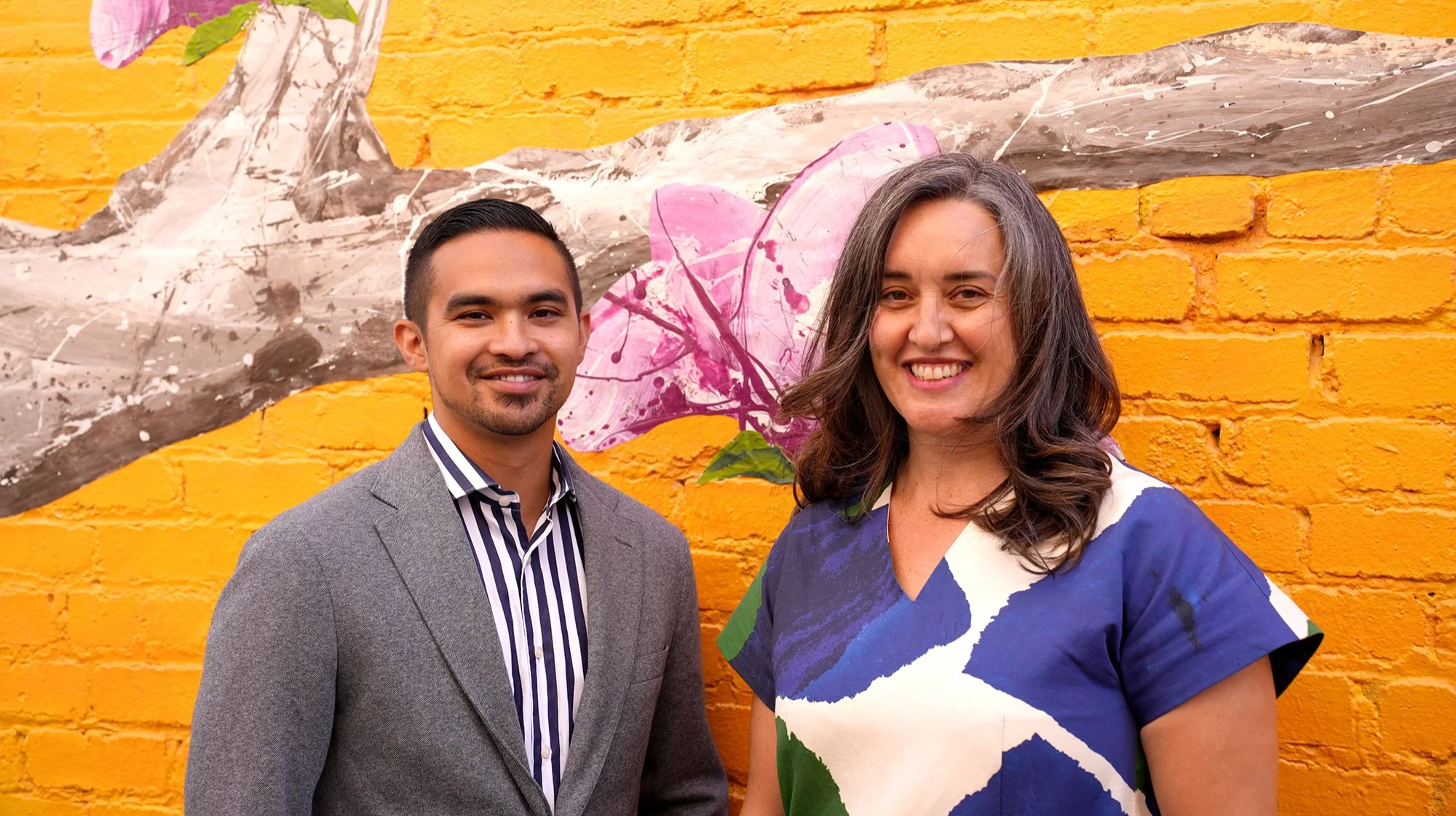 A man in a gray blazer with a striped shirt and a woman in a colorful dress standing in front of a yellow brick wall with a pink and gray mural.