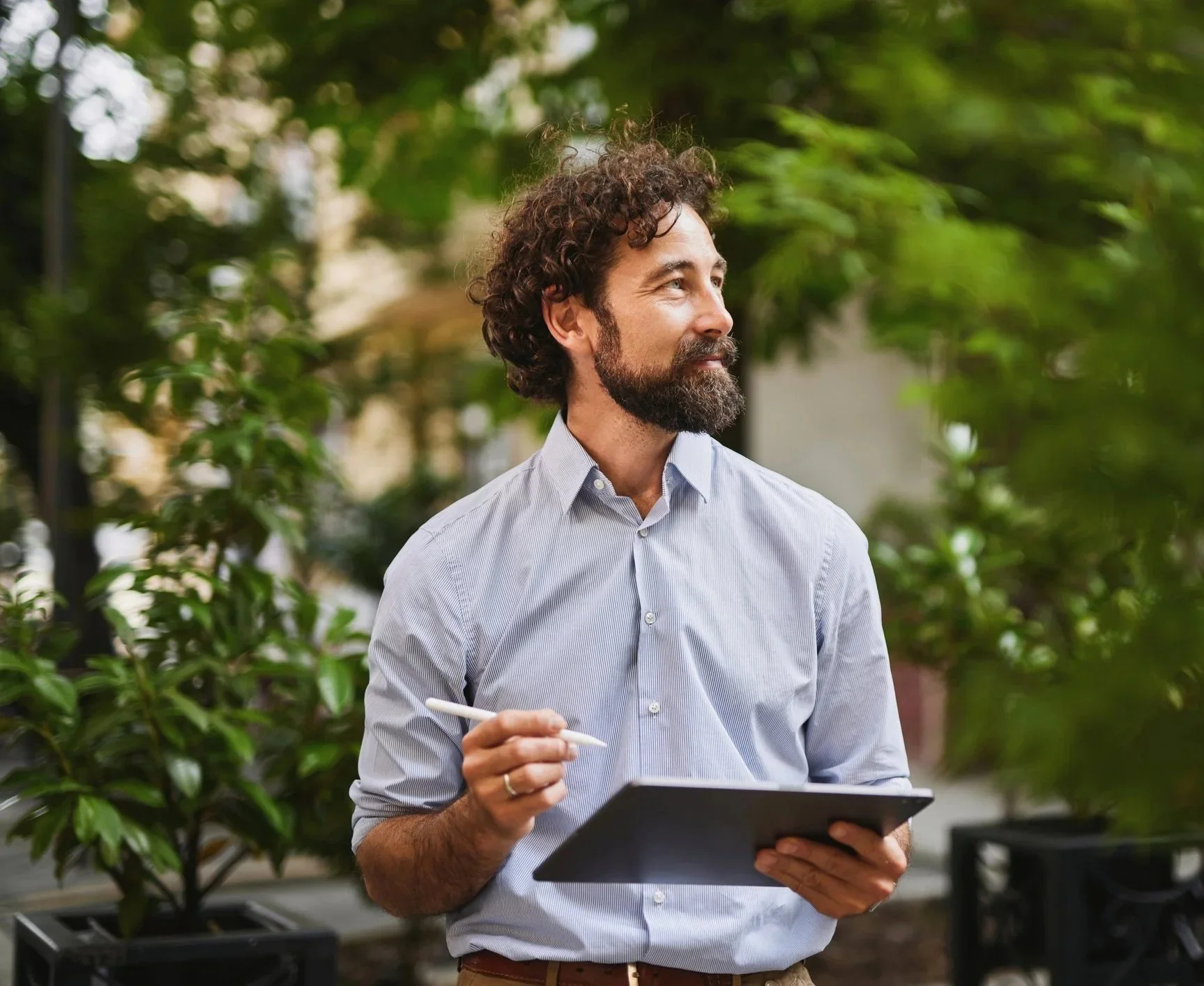 A man with curly hair and a beard, wearing a light blue button-up shirt, is standing outdoors amidst green trees and plants. He is holding a tablet in one hand and a stylus or pen in the other, looking to the right.