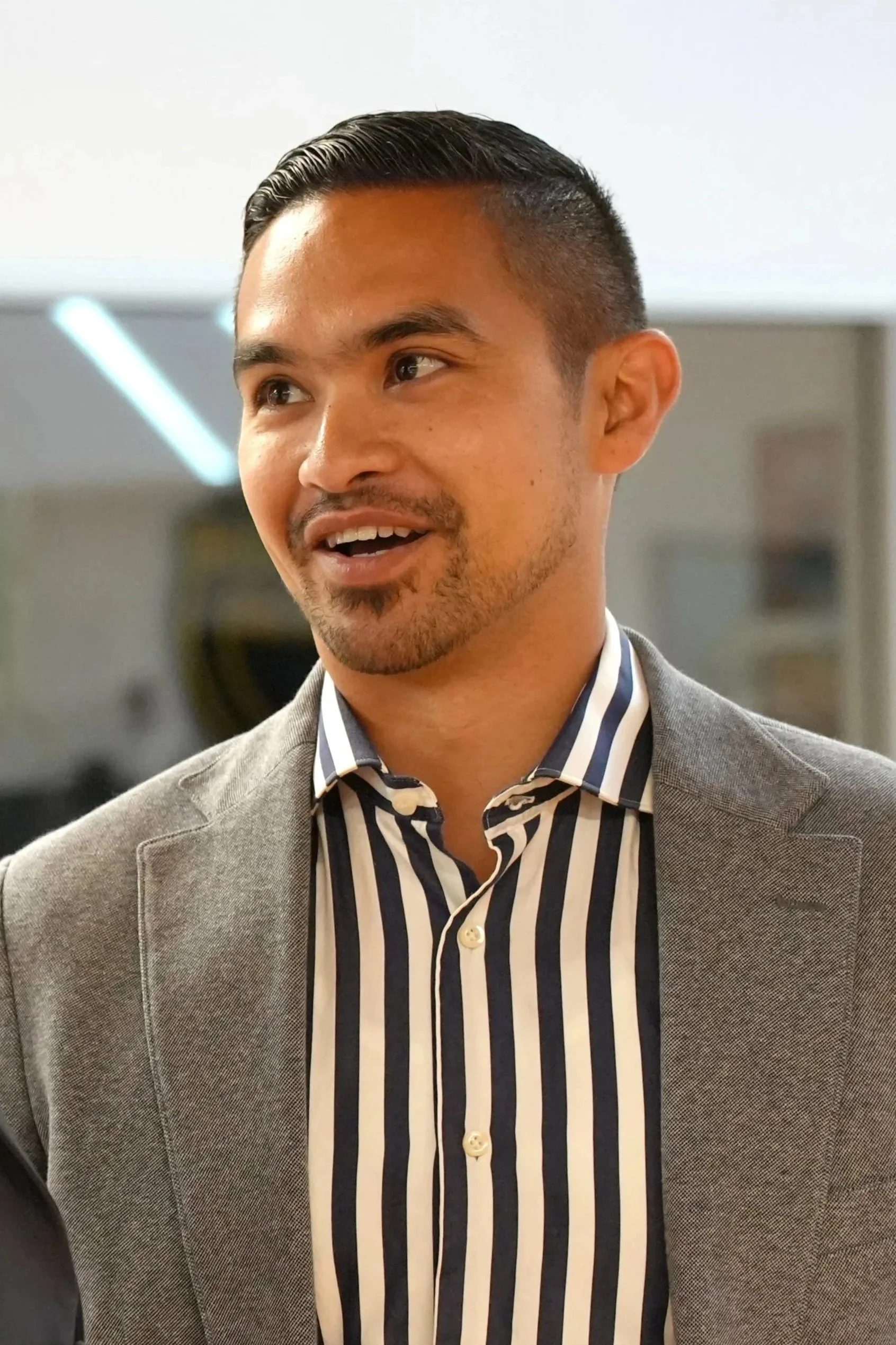 Close-up of a man with short black hair, wearing a light gray blazer and a black, white, and beige striped dress shirt, smiling and looking to the side, in an indoor setting.