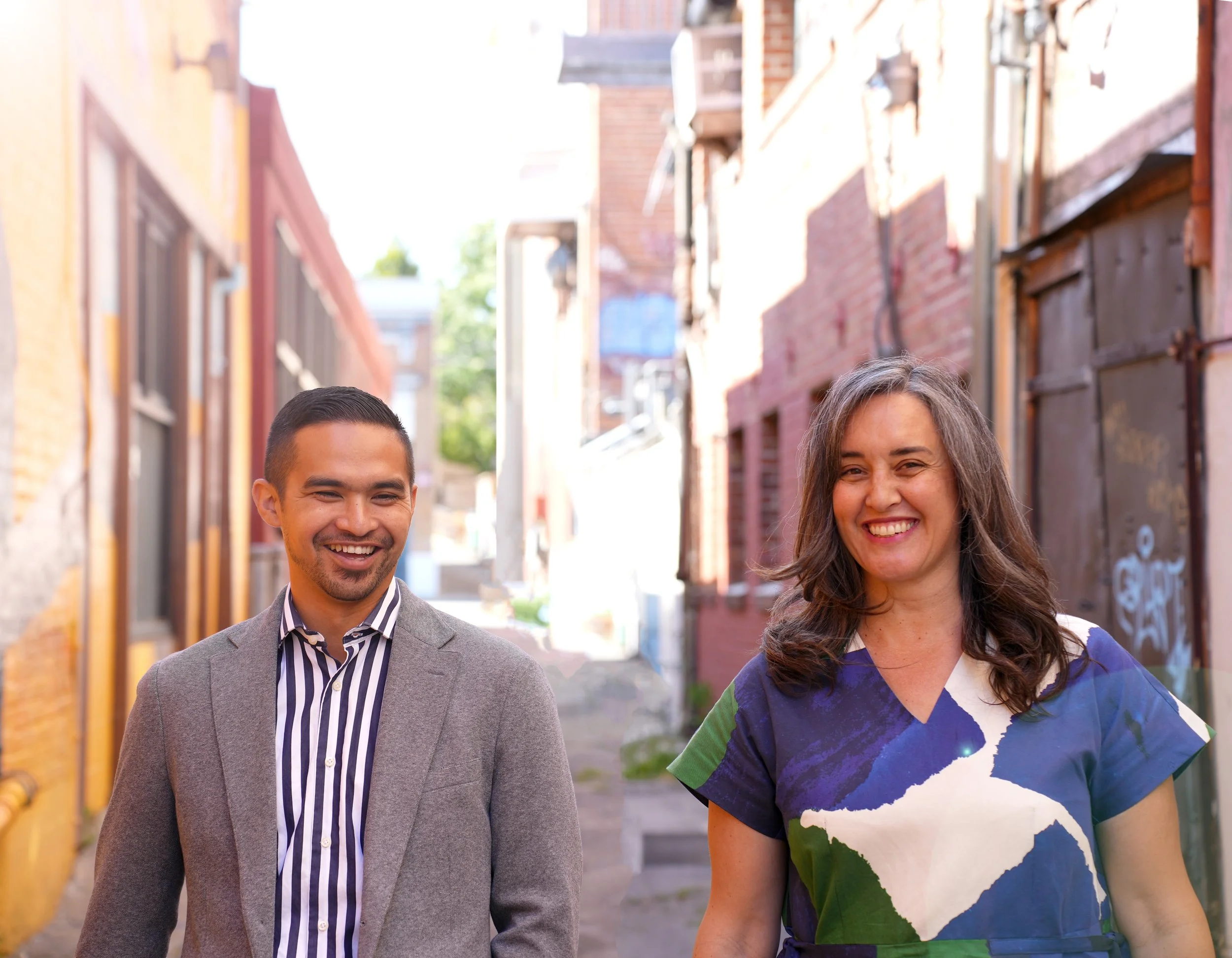 Two people, a man and a woman, smiling and walking outdoors in a narrow alley with colorful brick buildings.