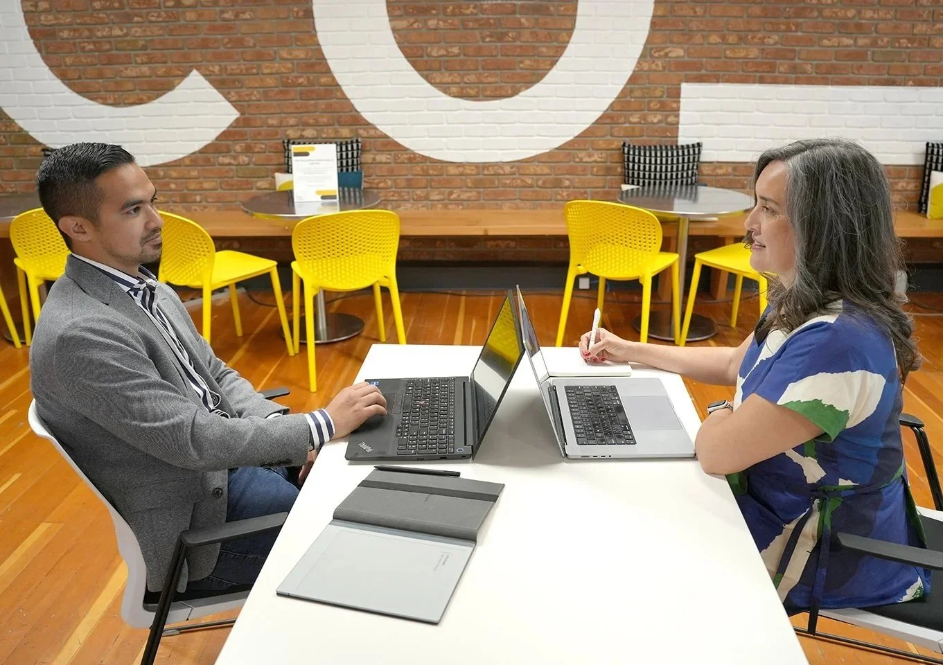 Two women sitting at a white table facing each other with laptops, in a modern office or cafe with yellow chairs and a brick wall background.
