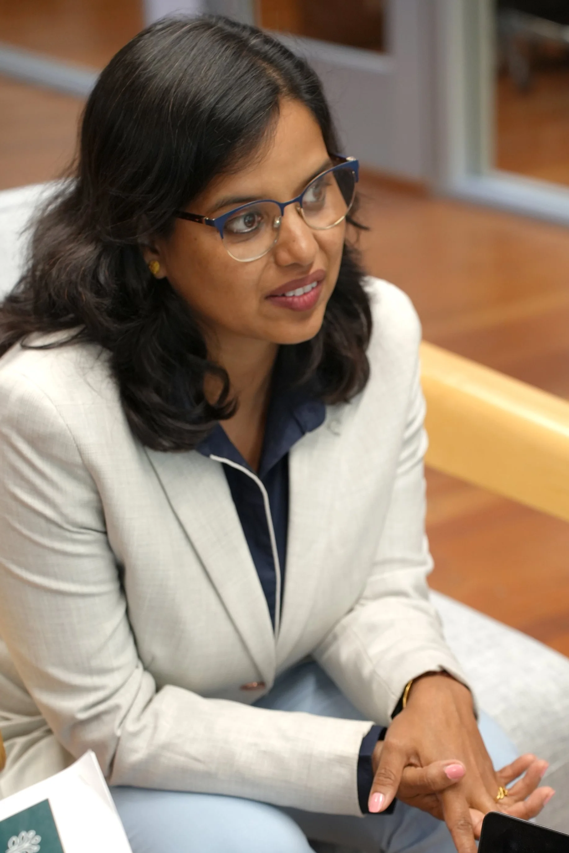 A woman with dark hair, glasses, and gold earrings, wearing a cream-colored blazer over a dark top, sitting at a table and holding hands with someone else's hand.