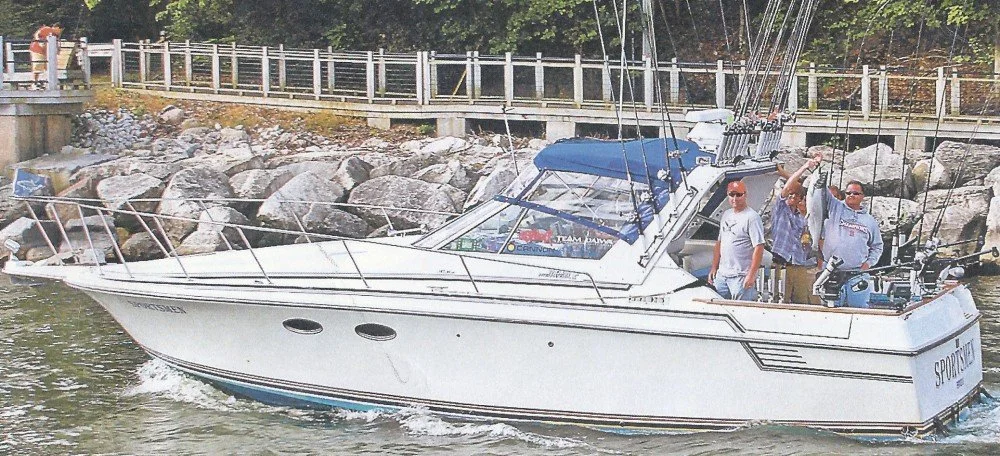A group of men on a white fishing boat with multiple fishing rods, docked near a rocky shoreline with a fence in the background.