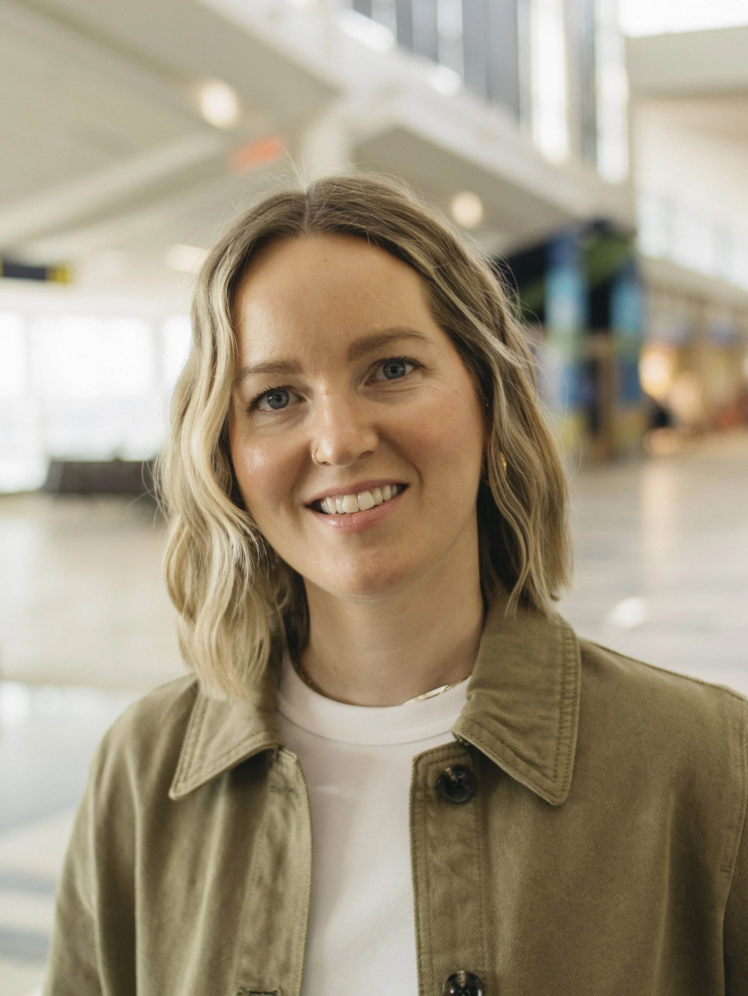 Close-up of a smiling woman with blonde wavy hair, wearing a tan jacket over a white top, in an indoor setting with blurred background.