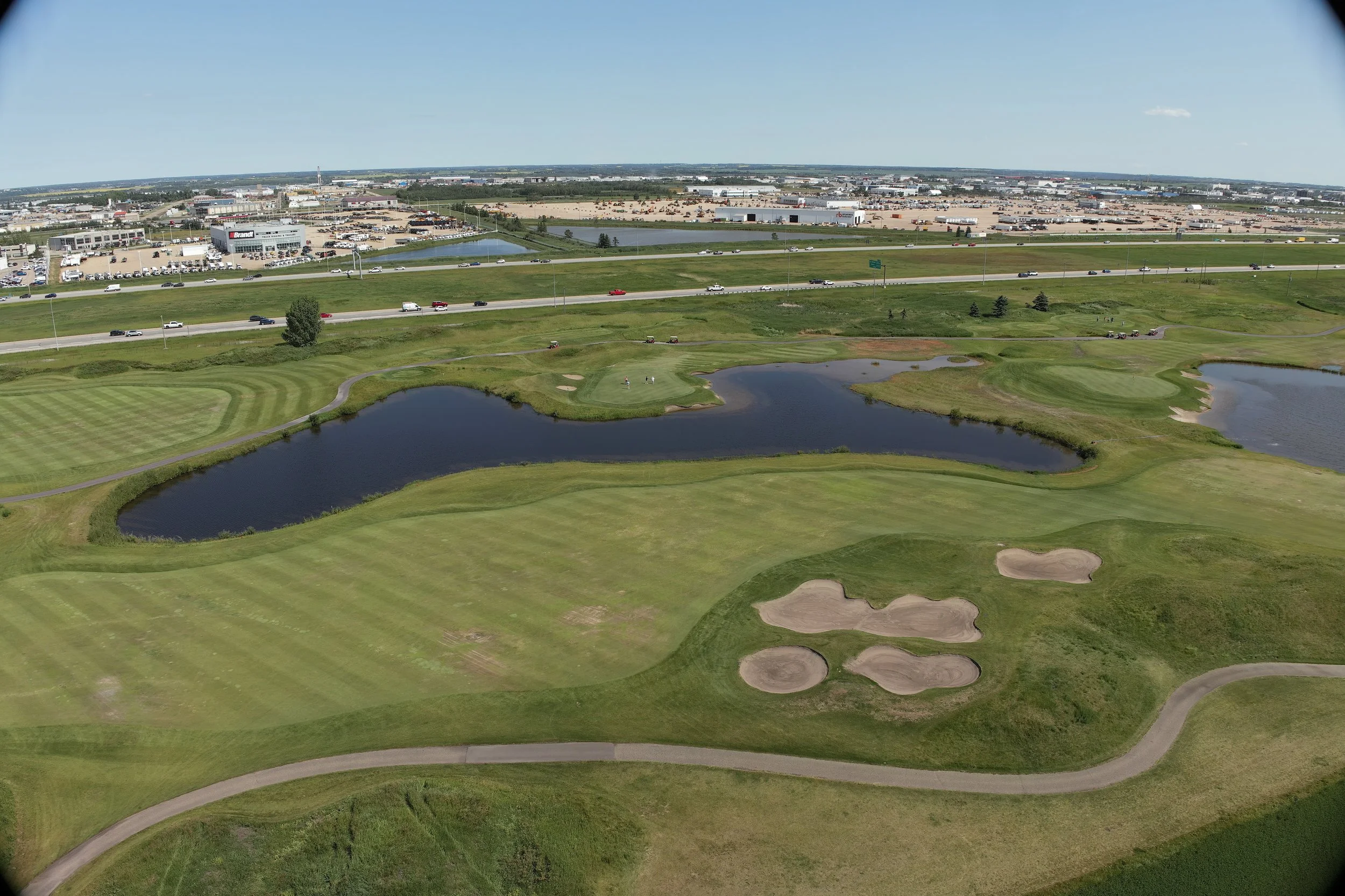 Aerial view of a golf course with water hazards and sand bunkers, adjacent to a highway and commercial retail area with parking lots and buildings.