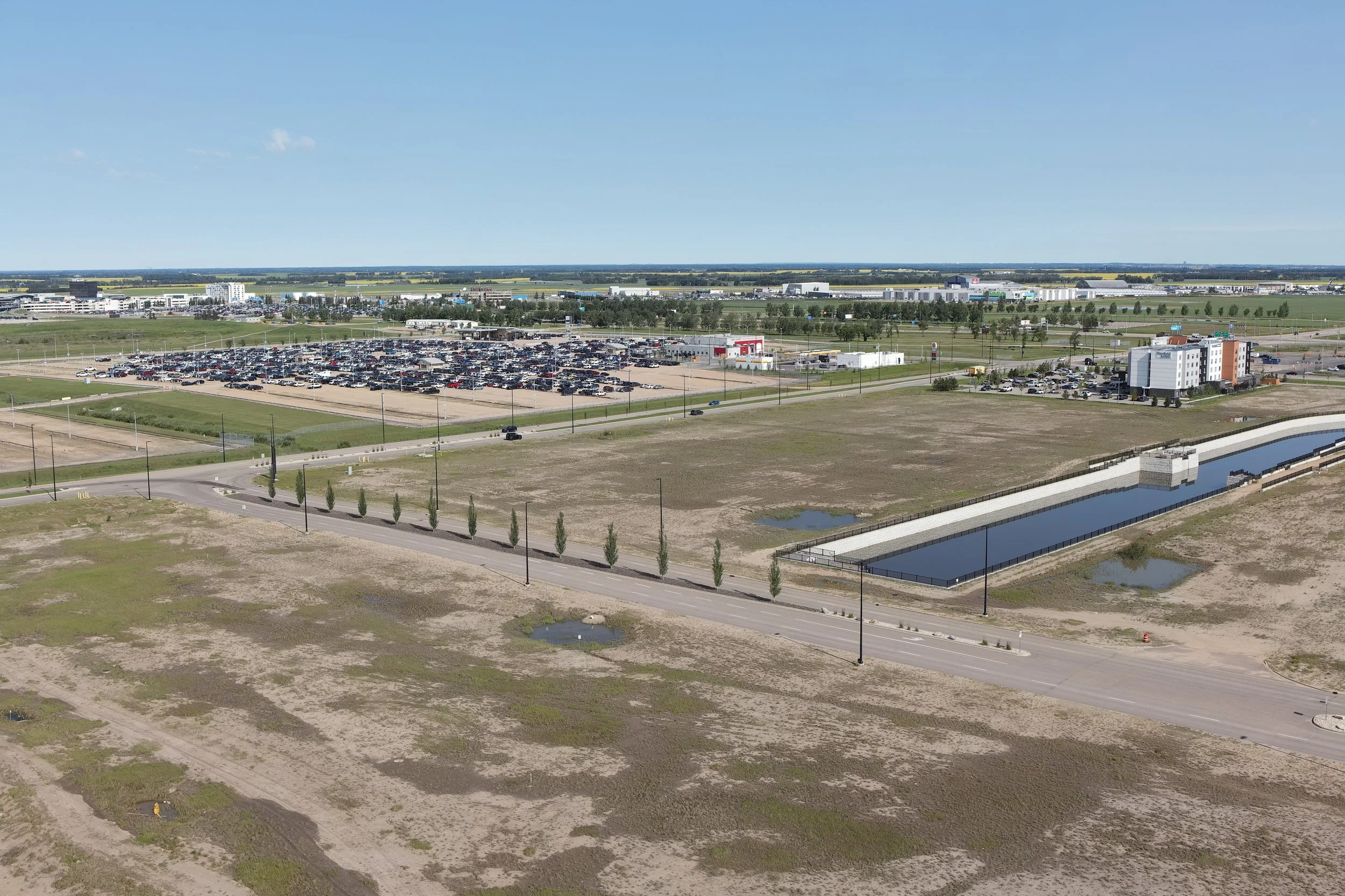 Aerial view of a large parking lot with many cars, nearby buildings, and open green spaces under a clear blue sky.