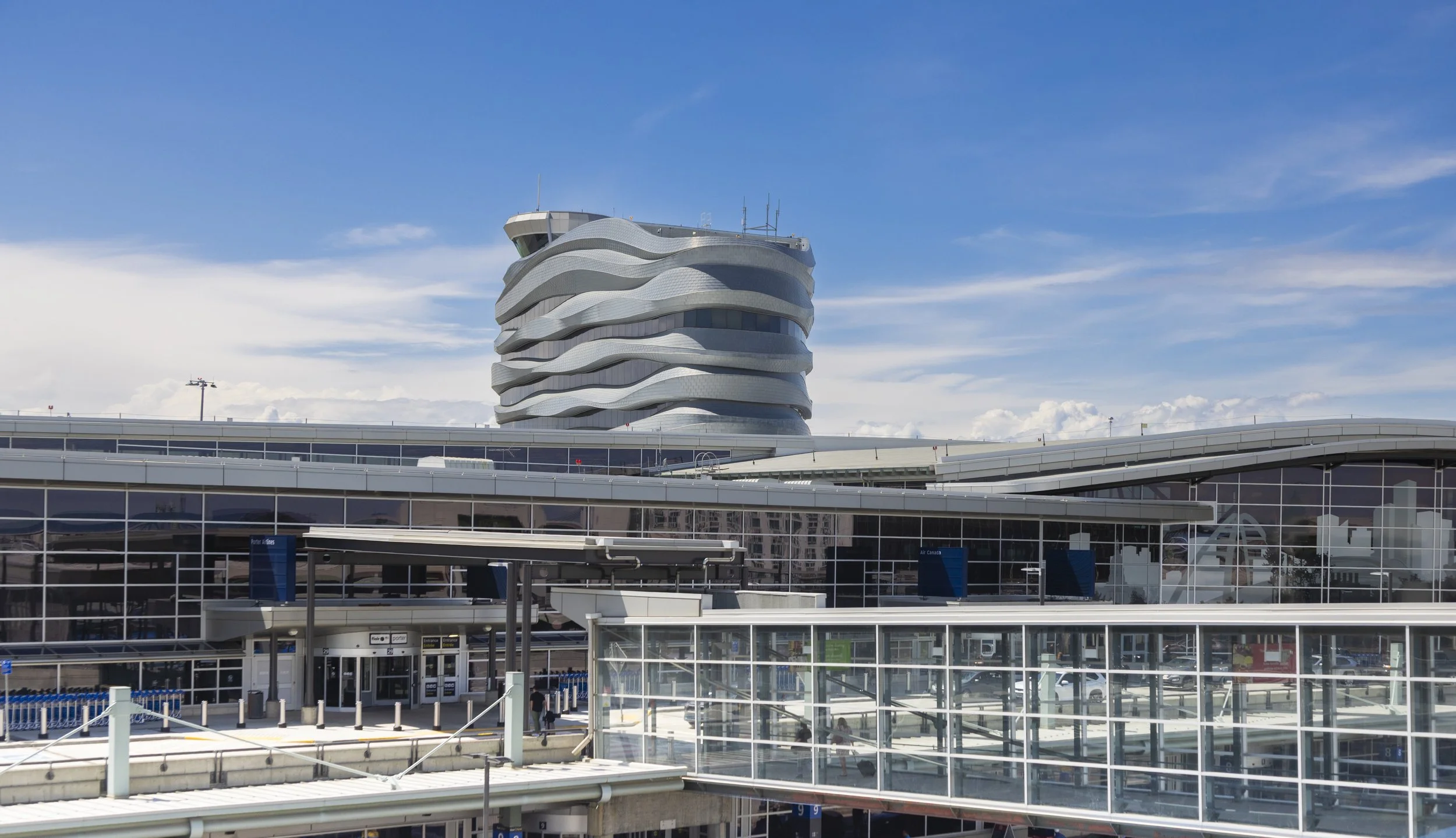 Modern airport terminal with a distinctive multi-wavy shaped building in the background, blue sky, and four floors visible.