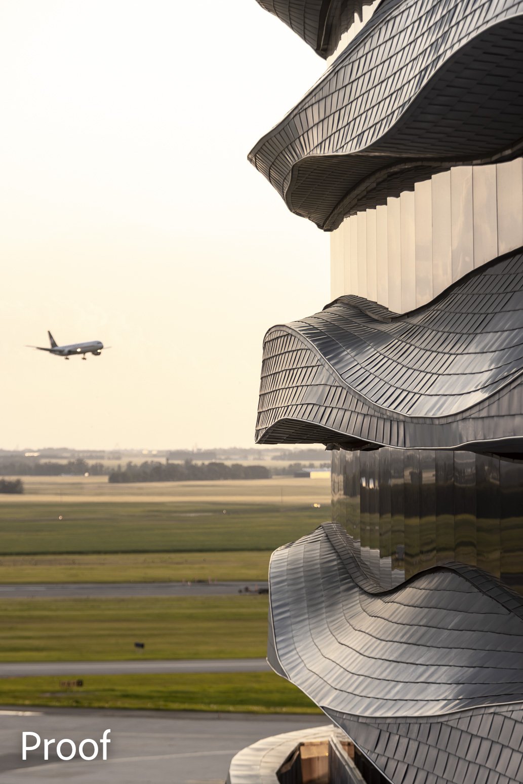 A modern building with a wavy metallic exterior reflecting light, adjacent to a flat open landscape with an airplane flying in the sky during sunset or sunrise.