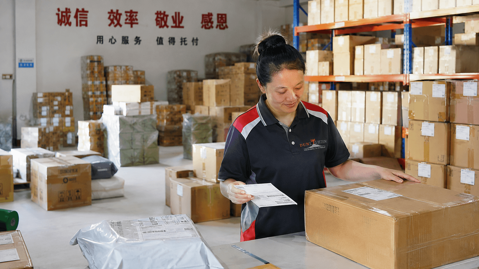 A woman in a black, red, and white uniform is inspecting a cardboard box in a warehouse filled with shelves of packed boxes.
