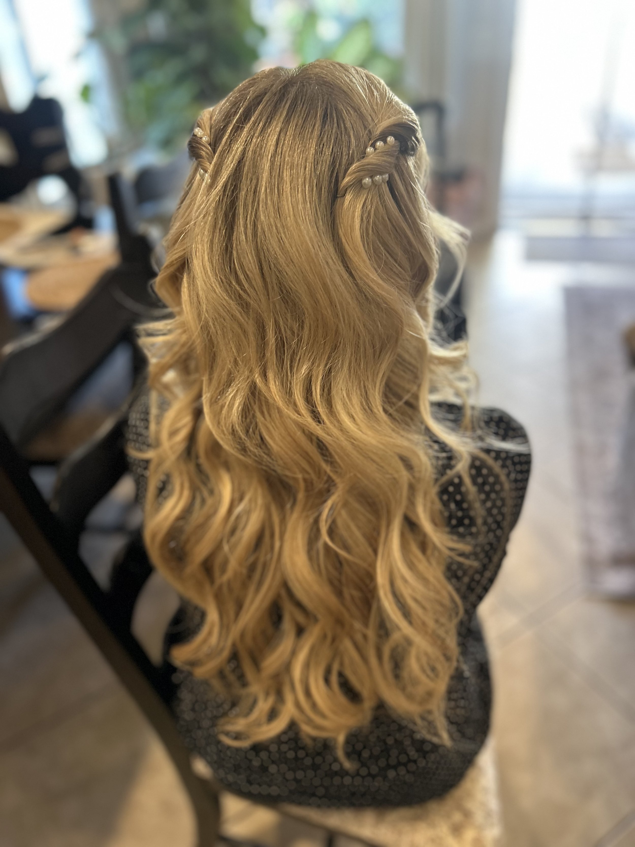 A woman with long, wavy, blonde hair styled in loose curls and small pearl clips, sitting in a salon or home setting.