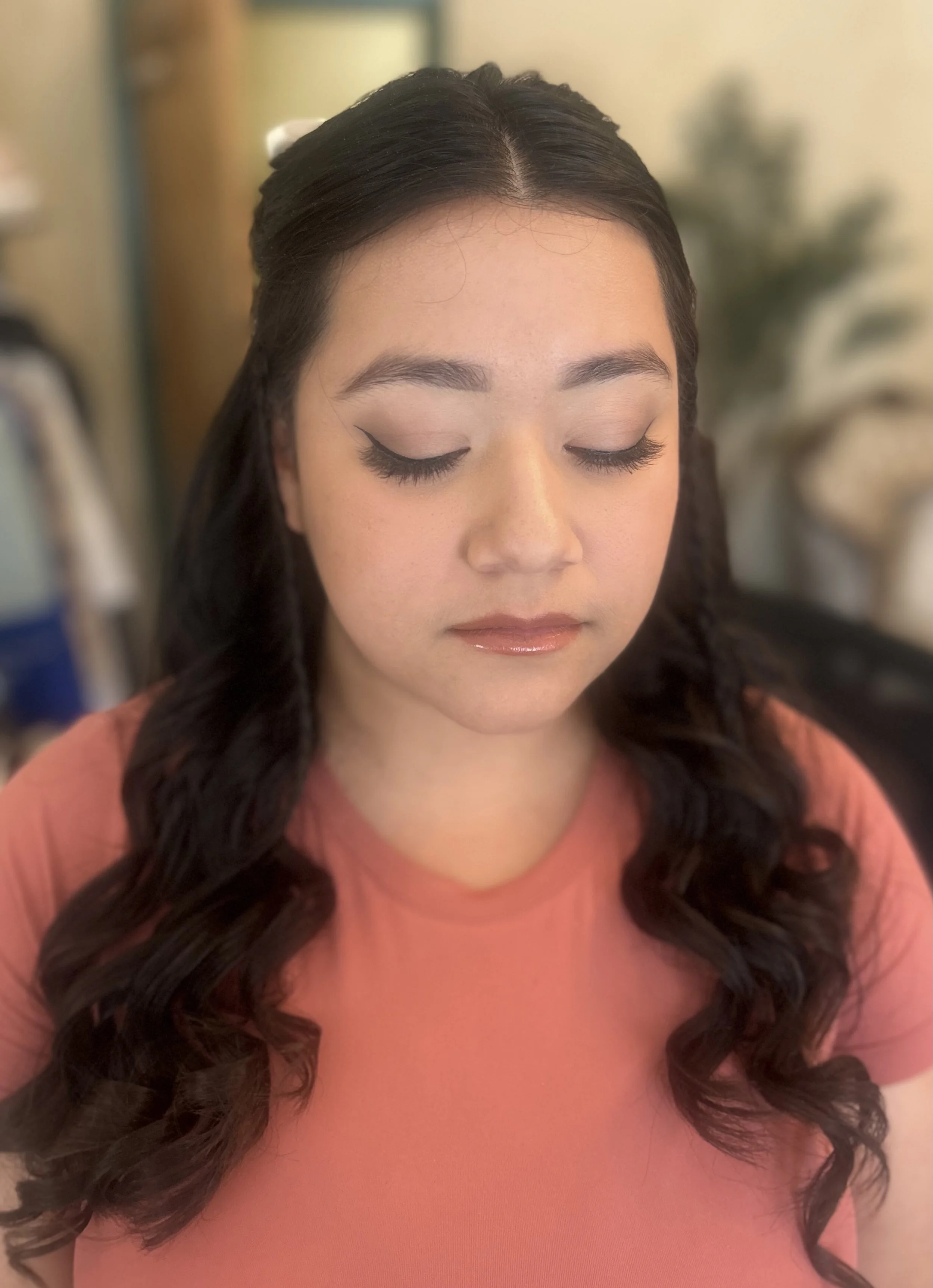 A woman with dark, wavy hair and makeup with winged eyeliner, closed eyes, and glossy lips, wearing a pink or peach-colored top.