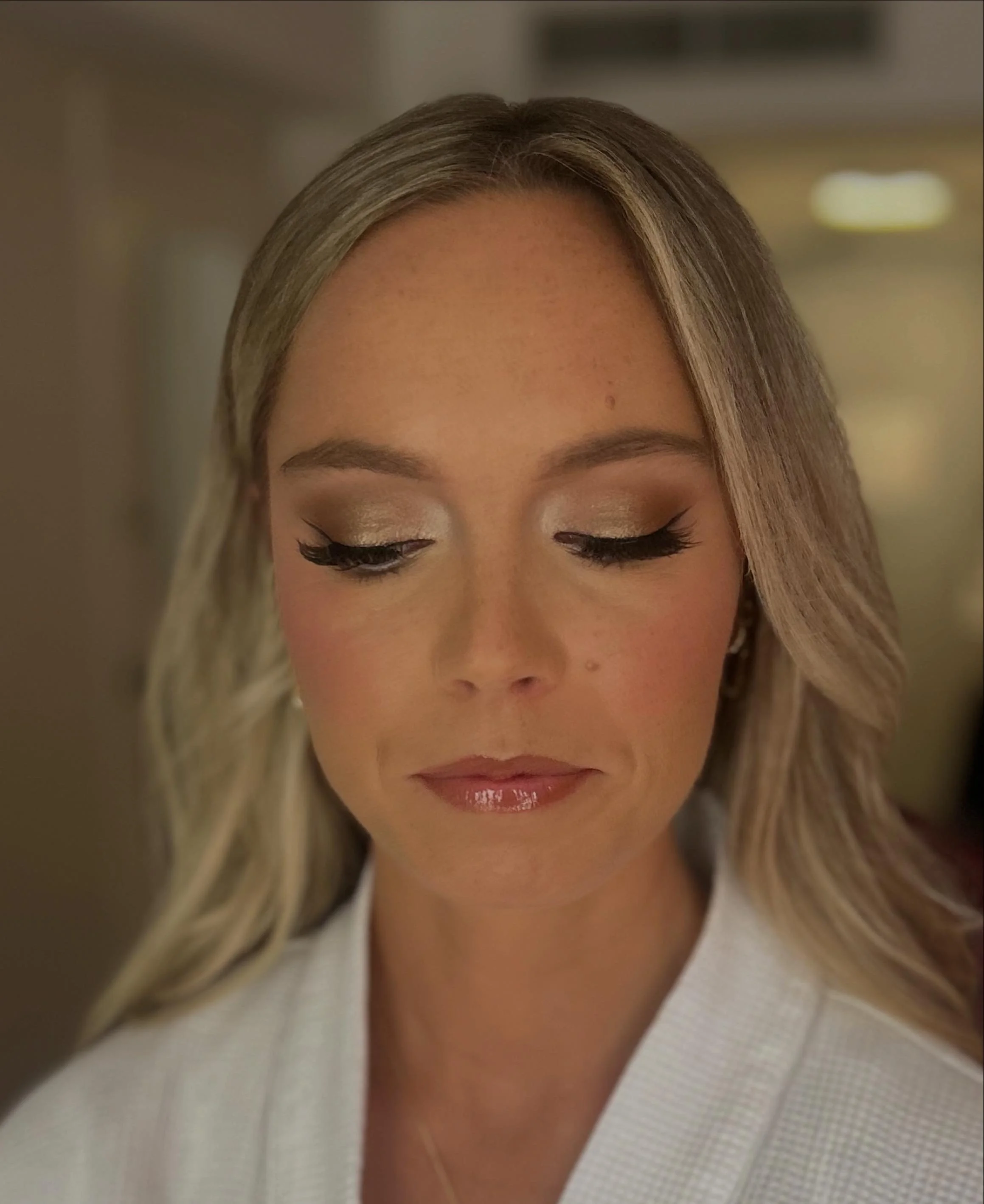 Close-up of a woman with blonde hair, makeup, and closed eyes, wearing a white textured robe.