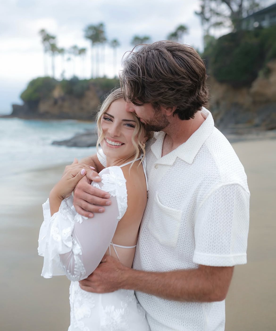 A smiling young woman and a man hugging on a beach, with palm trees and cliffs in the background.
