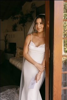 A woman in a white wedding dress standing near a doorway indoors, smiling and looking outside.