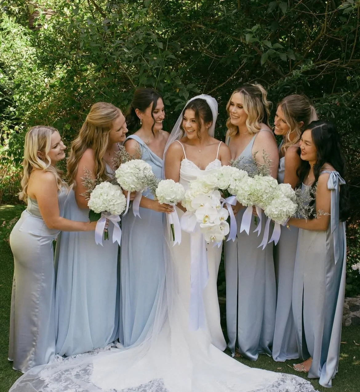 Bride and bridesmaids standing outdoors, smiling, holding white bouquets, dressed in light blue and white dresses, with greenery background.