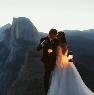 A couple dressed in formal attire, holding lanterns, standing on a rocky ledge with a mountain landscape in the background at dusk or dawn.