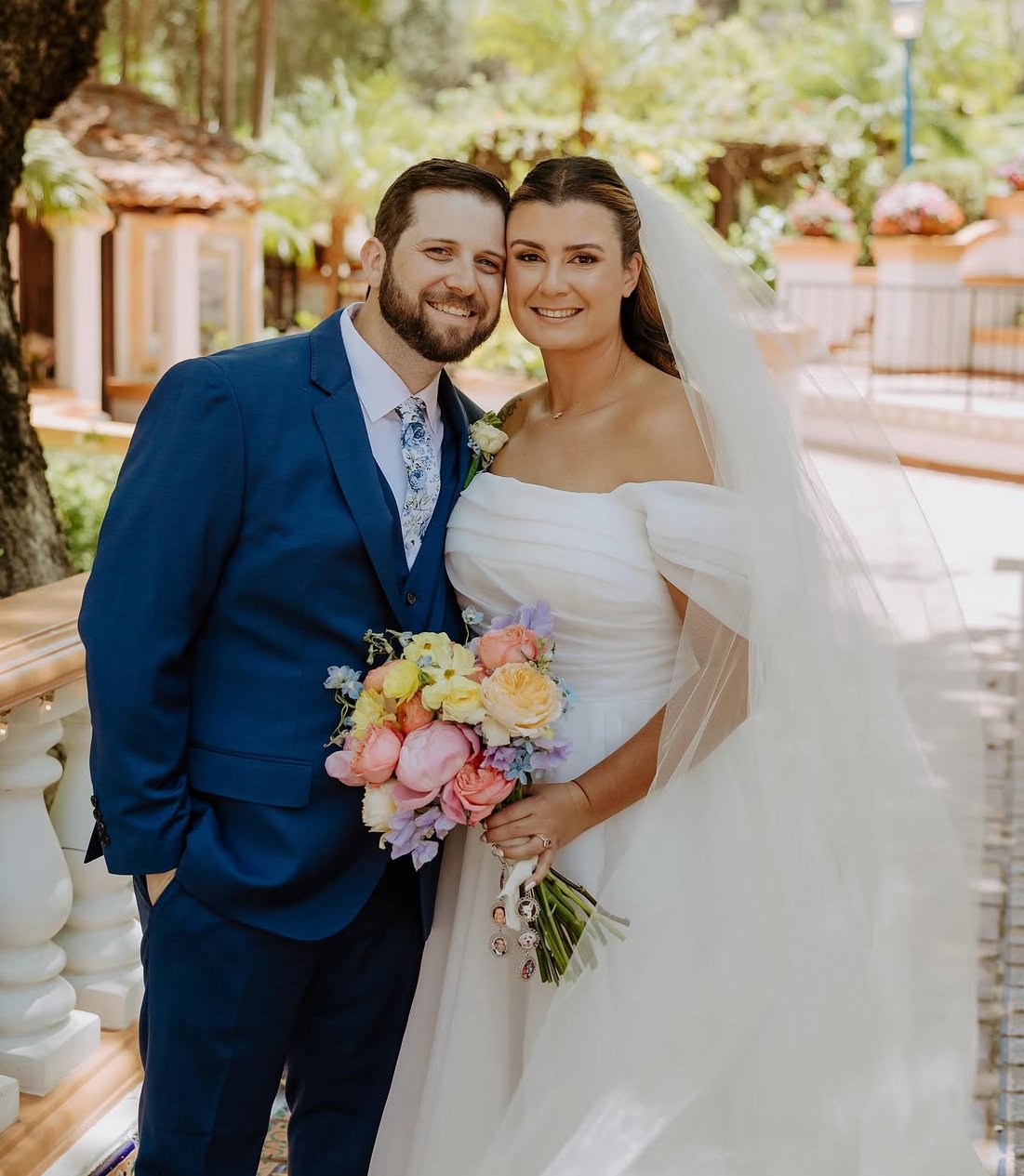 Happy bride and groom posing outdoors on wedding day, woman in white wedding gown holding a bouquet of flowers, man in blue suit with floral tie.