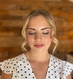 A woman with blonde hair styled in loose curls, wearing a white polka dot top, with her eyes closed, smiling softly in front of a wooden background.