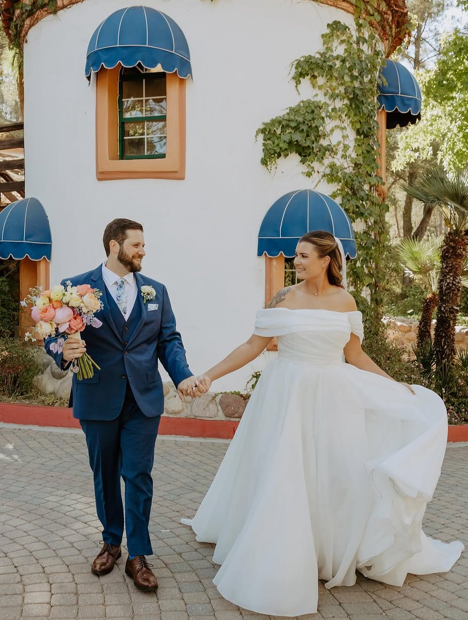 A bride and groom holding hands and smiling outside a building with blue awnings, surrounded by trees and plants.