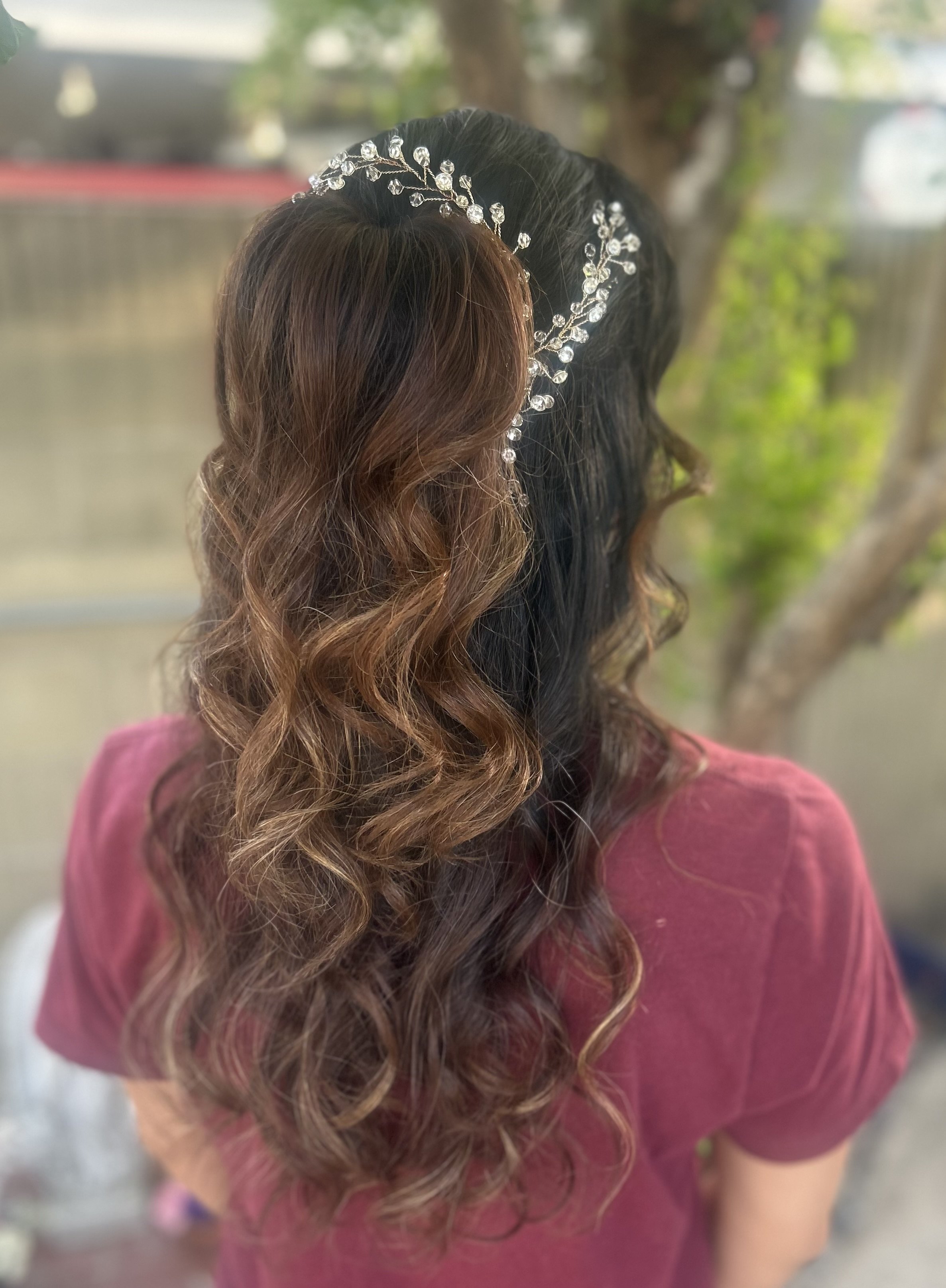 The back of a person with long, wavy hair styled in loose curls, wearing a silver headpiece with decorative beads, and a pink shirt outdoors.