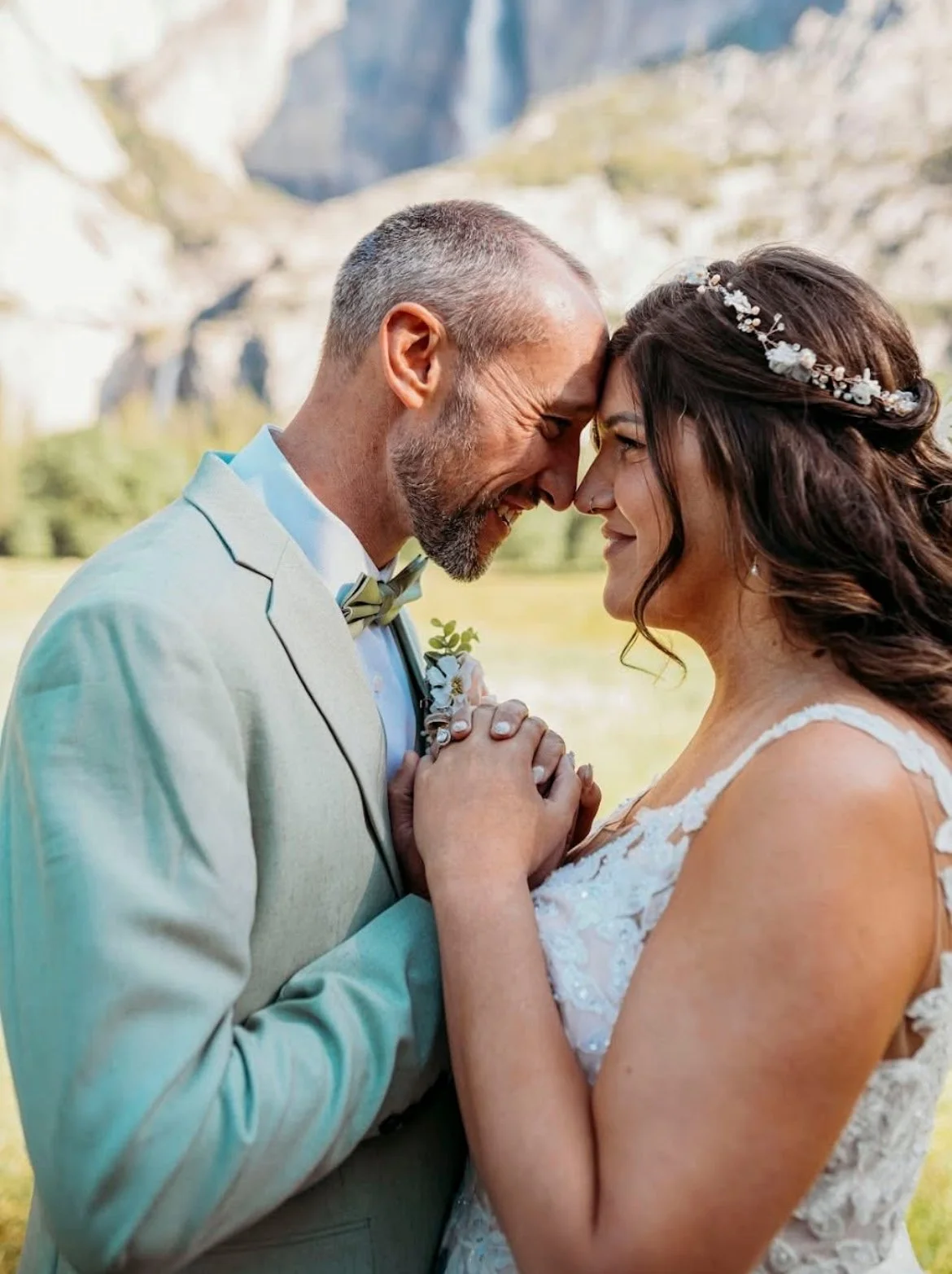 A couple in wedding attire touching foreheads and holding hands outdoors with mountains in the background.
