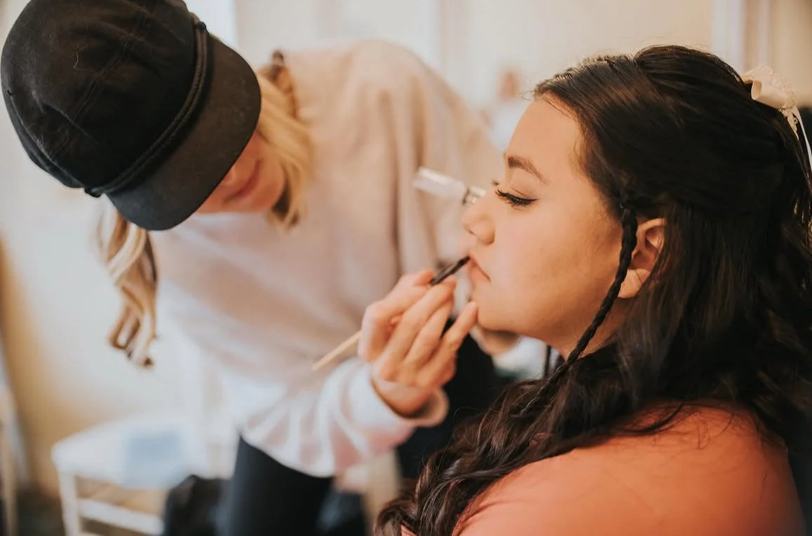 A woman with long dark hair getting her makeup done, with another woman applying makeup near her lips.