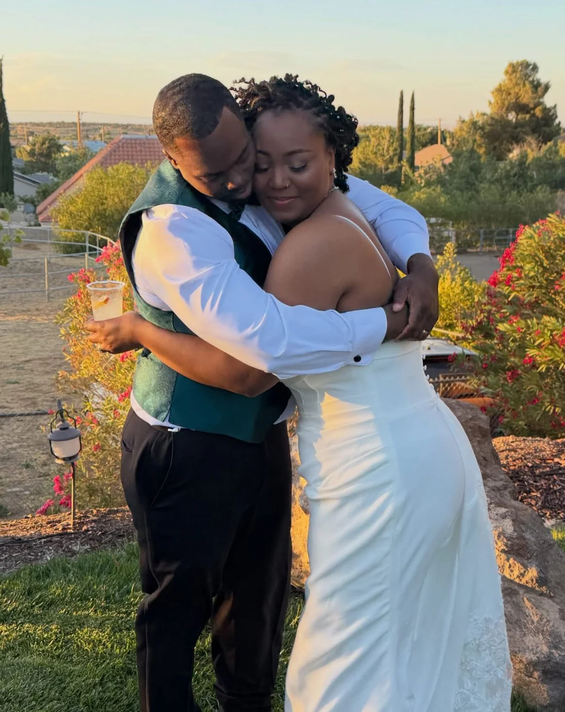A couple embraces outdoors during sunset, with the woman wearing a white dress and the man in a vest and dress shirt, holding a drink.