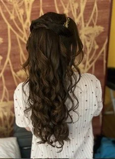 Woman with long, wavy brown hair and a small hair accessory, facing away, wearing a white shirt with small black dots, in front of a decorative background.