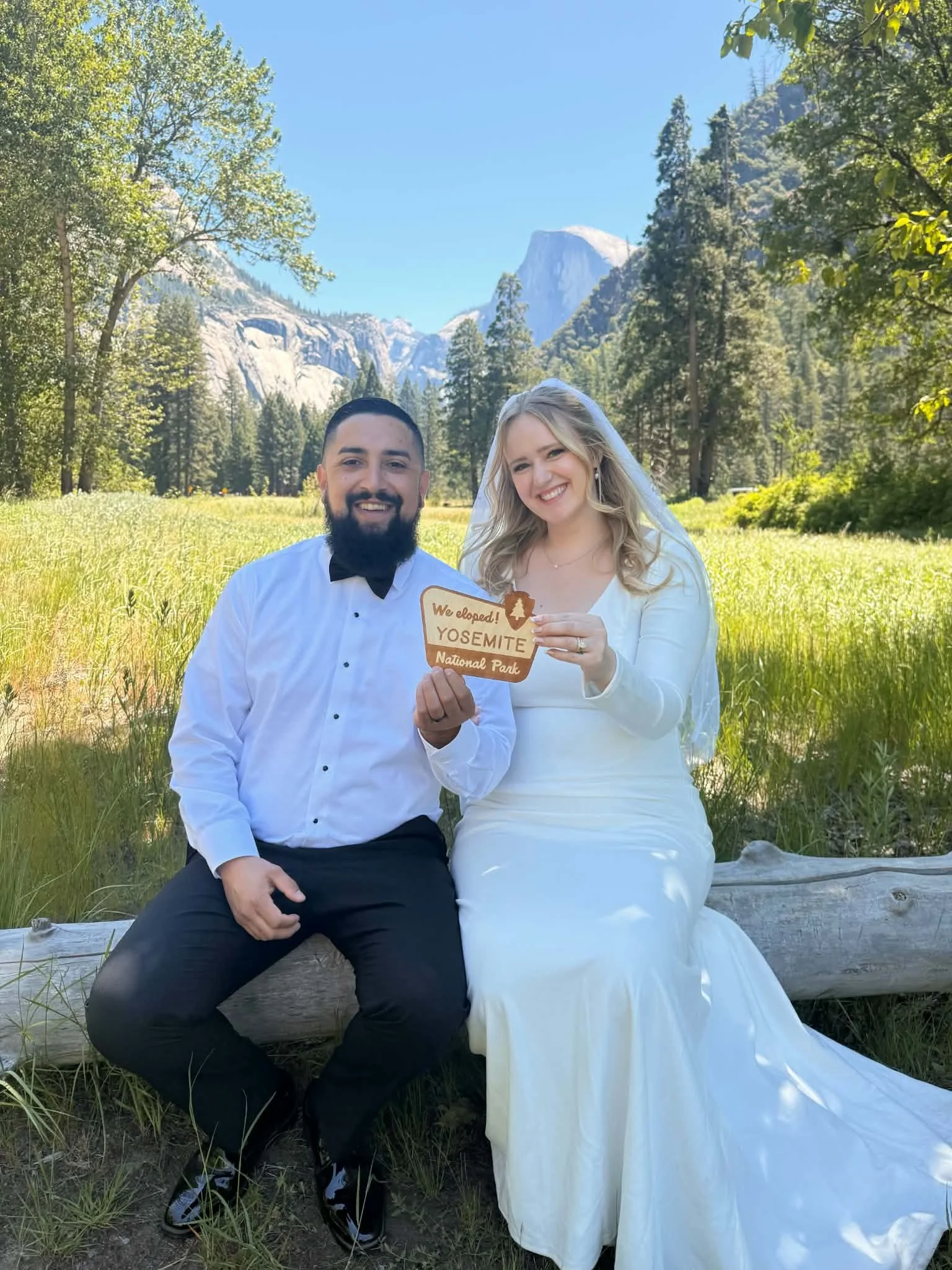 A wedding couple dressed in wedding attire sitting on a log in a forested area of Yosemite National Park, holding a wooden sign that reads 'Weelope! Yosemite National Park' with mountains and trees in the background.