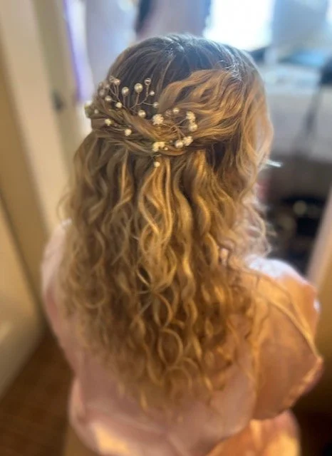 A woman with long, curly blonde hair wearing a decorative hairpiece with small white flowers and pearls, viewed from behind.
