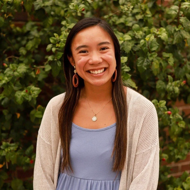 A young woman smiling outdoors in front of green bushes, wearing a blue dress, beige cardigan, and jewelry.