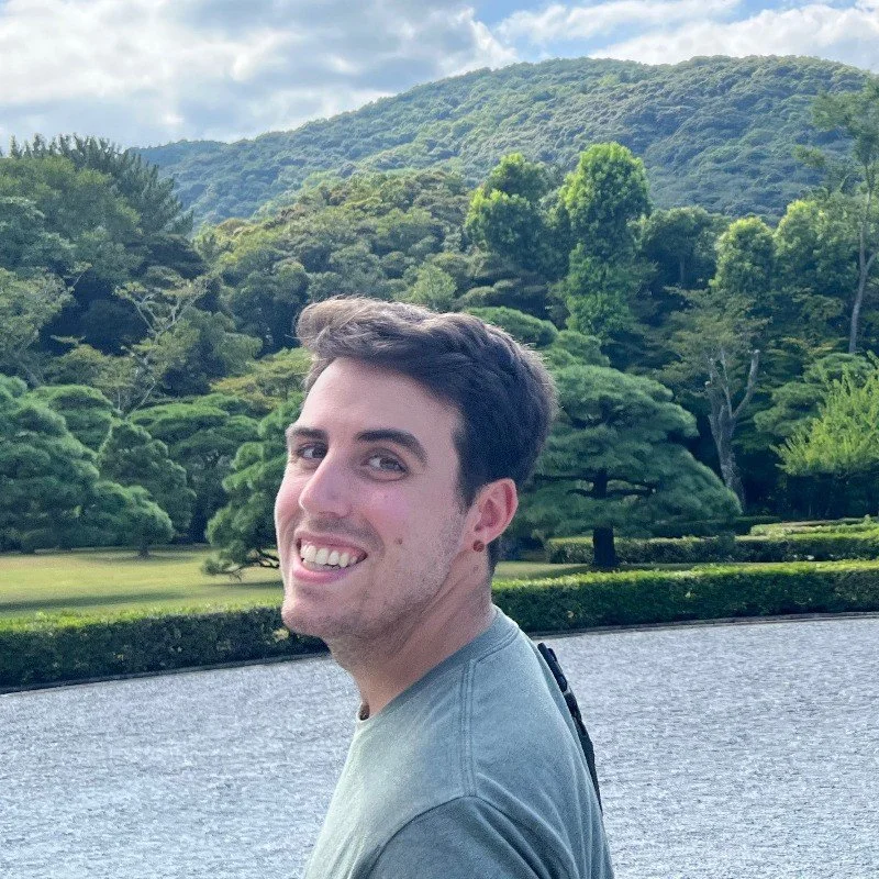 Young man smiling outdoors by a body of water with lush green trees and mountains in the background.