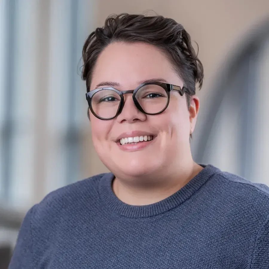 A young person with short brown hair, glasses, and a big smile wearing a blue sweater, in an indoor setting with blurred background.