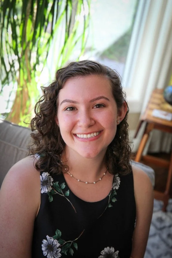 A young woman with curly brown hair smiling, wearing a sleeveless black dress with a floral pattern and a pearl necklace, sitting in a room with a window and green plants in the background.